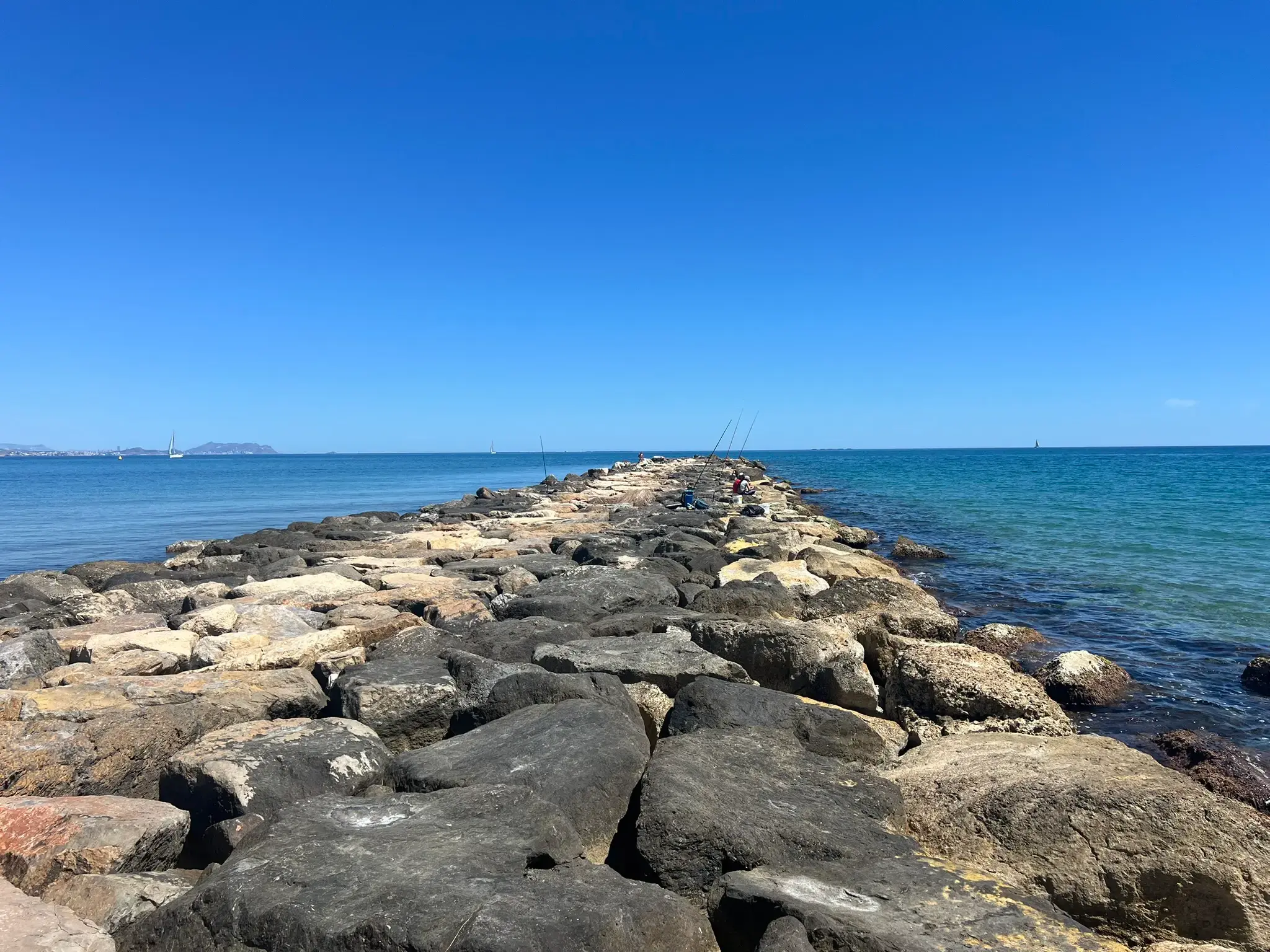 A long rock breakwater structure at Platja del Carrer la Mar in El Campello on a bright, sunny day. The camera looks down the center of the large grey and brown boulders extending into the calm, turquoise Mediterranean Sea. Several people are fishing with long rods near the end, and a single sailboat is visible in the distant water under a clear blue sky.