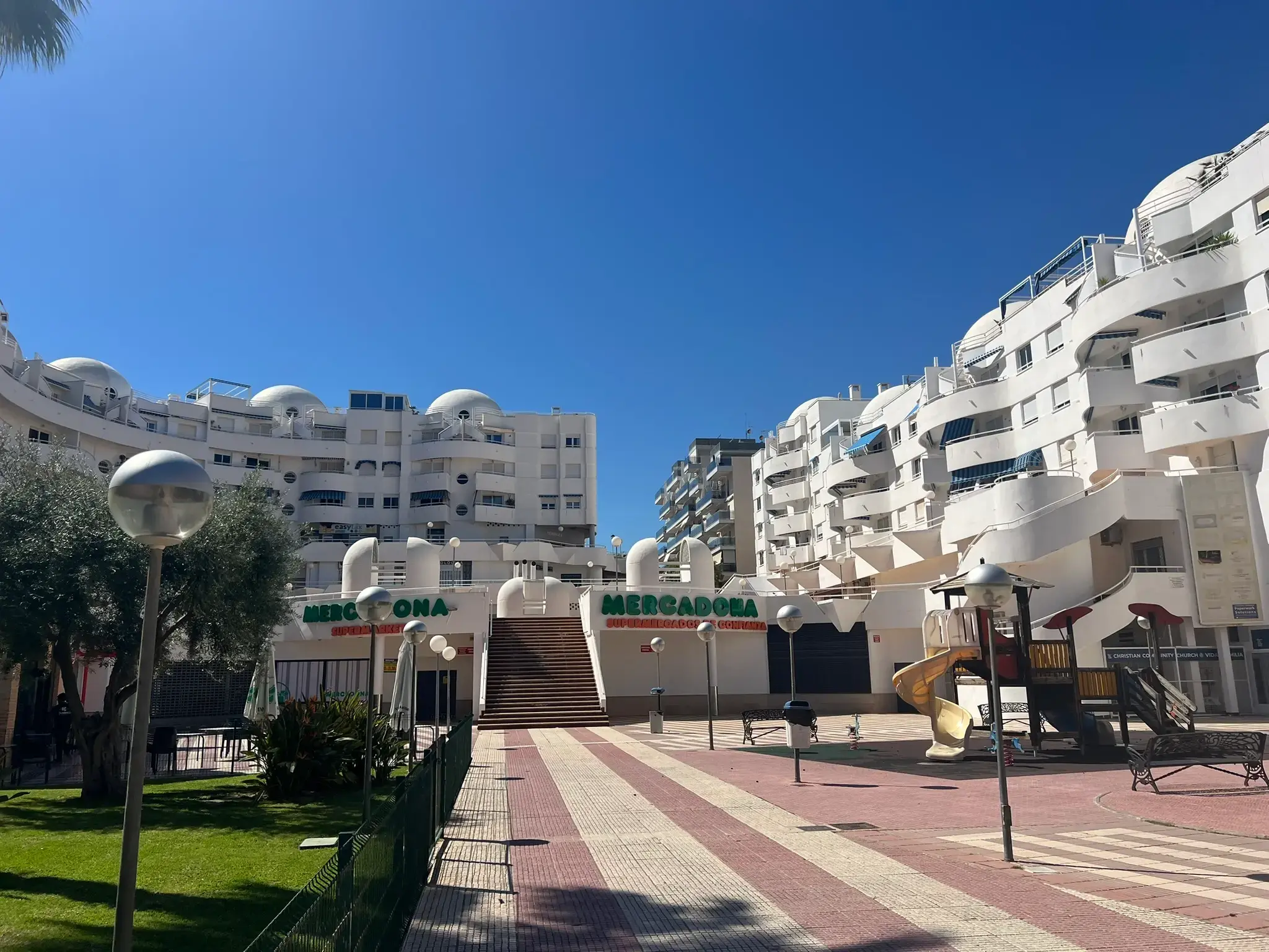 A sunny urban square in El Campello featuring a white multi-story residential complex with unique rounded balconies. In the center is a Mercadona supermarket entrance with a large staircase, a children's playground to the right, and a paved walkway with street lamps.