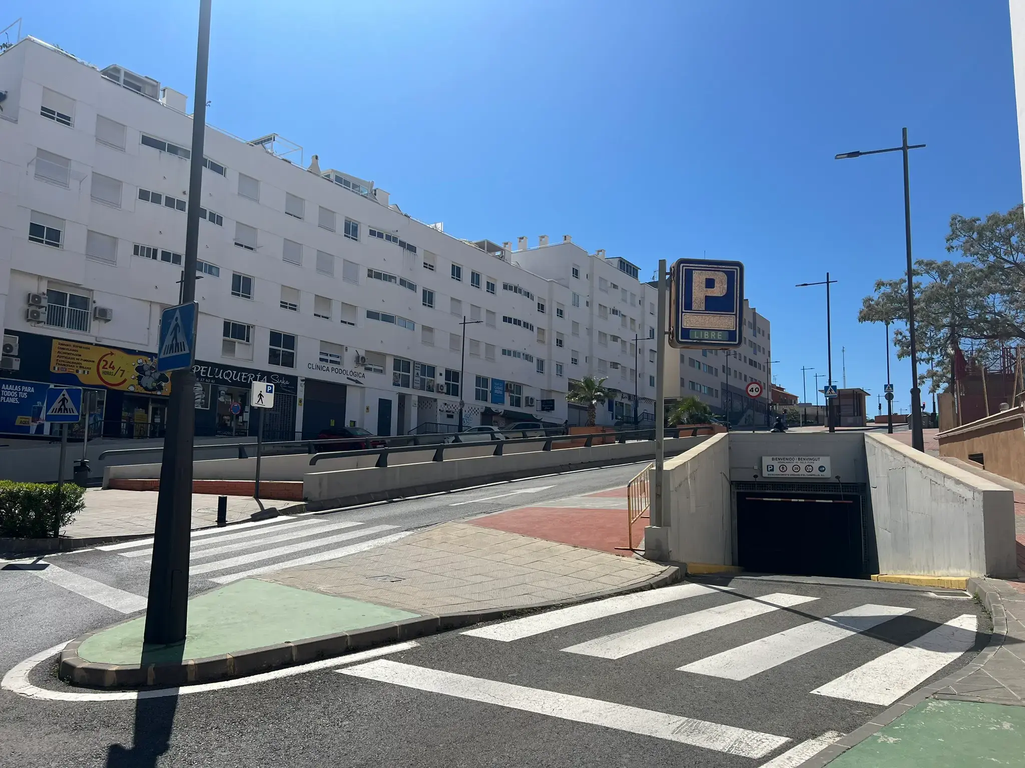 A bright street scene in El Campello showing the entrance to an underground parking garage with a P sign. The area includes white multi-story apartment buildings, several pedestrian crosswalks, and retail shops under a clear, sunny sky.