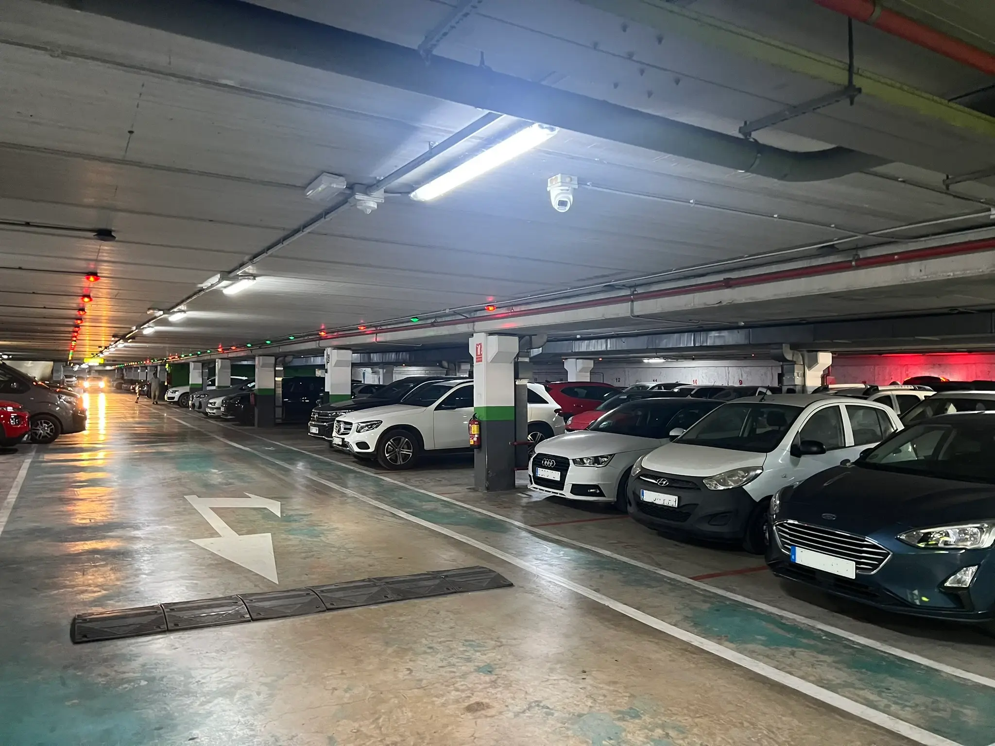 Interior view of a brightly lit underground parking garage in El Campello. Rows of various cars are parked in numbered spots, and a white directional arrow is painted on the floor.