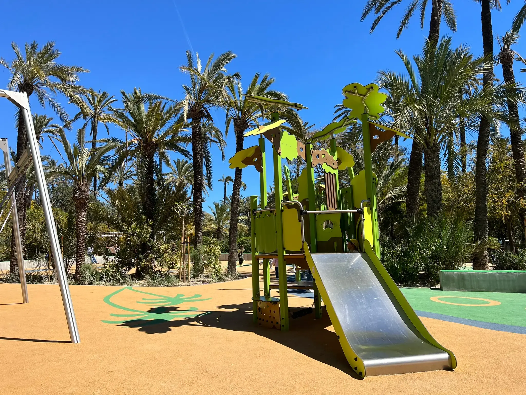 Children's playground at El Palmeral Park in Alicante with slides and climbing frames set in a shaded palm grove.