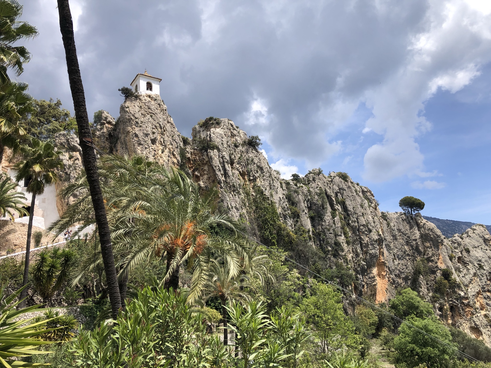 Der weiße Glockenturm des Castillo de San José auf einer Felsspitze über Guadalest, gesehen vom Palmengarten darunter unter bewölktem Himmel, mit einem kleinen Gebäude und Besuchern auf dem Weg am Fuß des Felsens