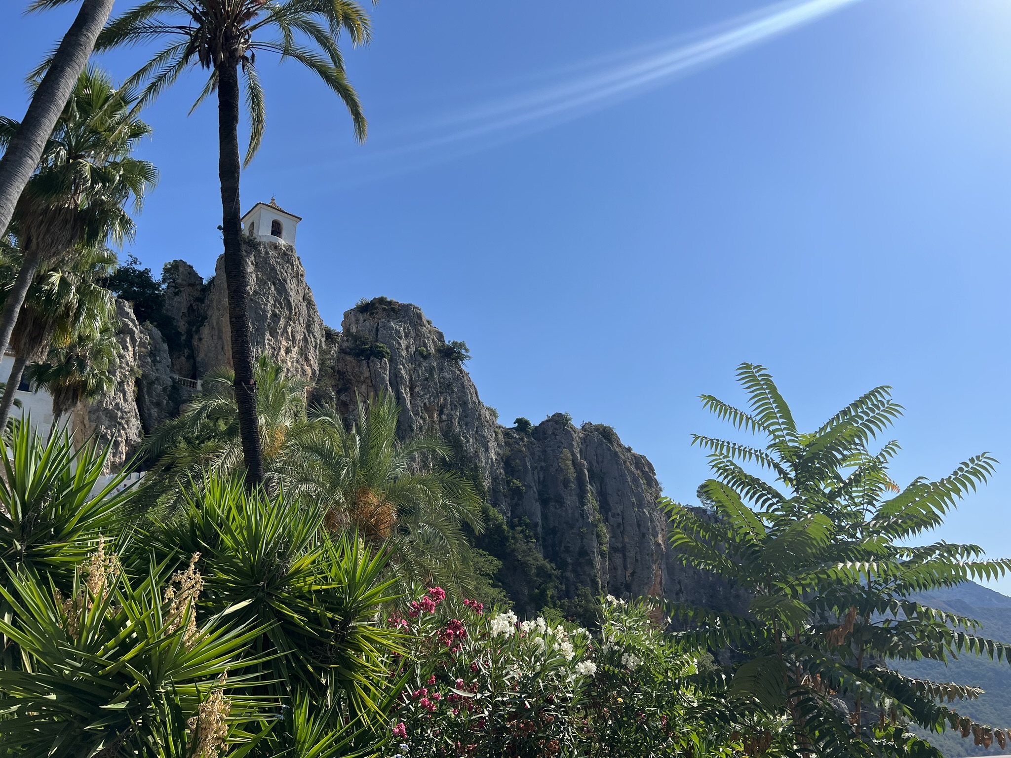 Der Glockenturm von Guadalest über dem Felsen, umrahmt von Palmwedeln sowie blühenden Oleander- und Yuccapflanzen im Vordergrund