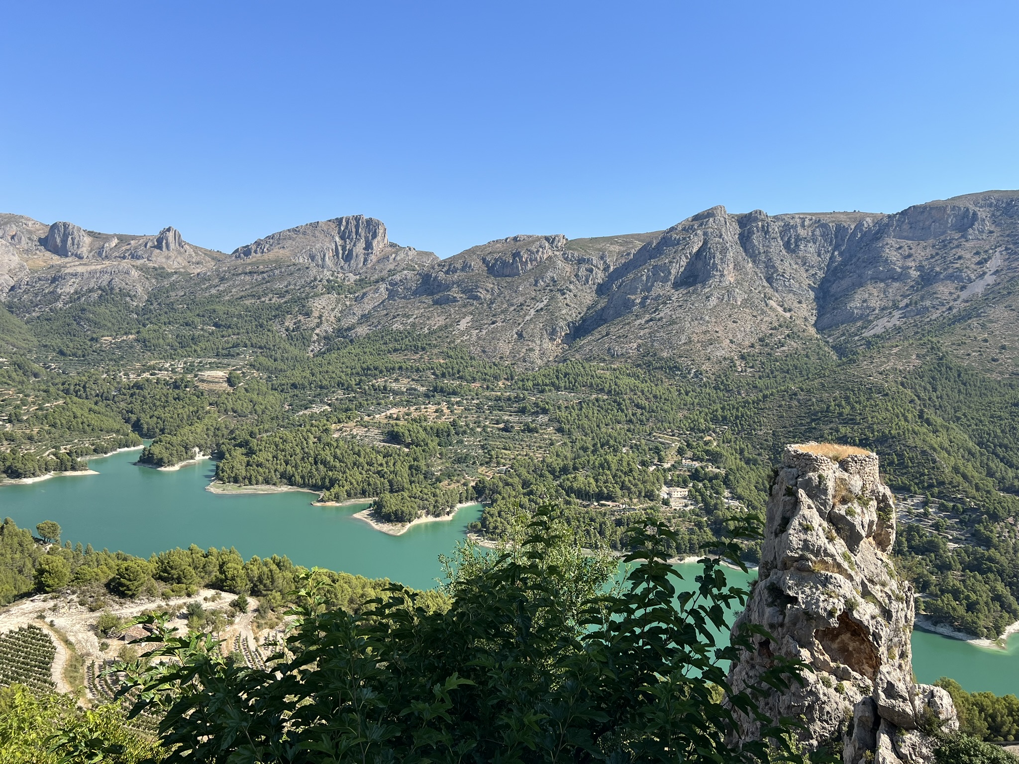 Der türkisfarbene Stausee Embalse de Guadalest von oben gesehen, mit einem Felsvorsprung und Burgruine im Vordergrund und terrassierten Feldern darunter