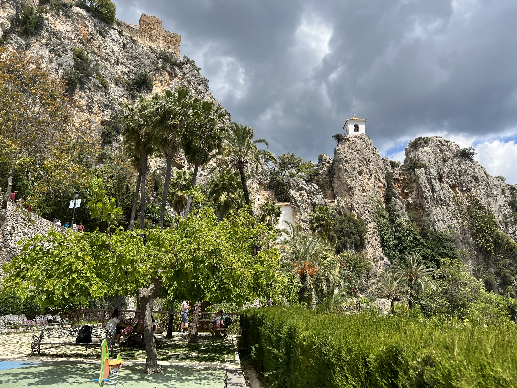 Ein Garten unterhalb des Burgfelsens von Guadalest mit Palmen, schmiedeeisernen Bänken und einem Kinderspielplatz, darüber die zerfallene Burgmauer und der Glockenturm unter stürmischem Himmel