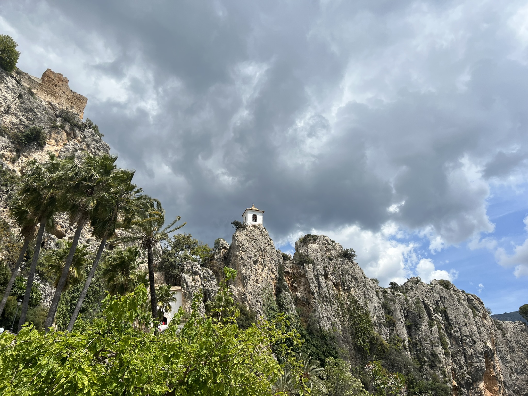 Die zerfallene Burgmauer und der Glockenturm auf der Felsspitze in Guadalest, umrahmt von hohen Palmen unter dramatisch bewölktem Himmel