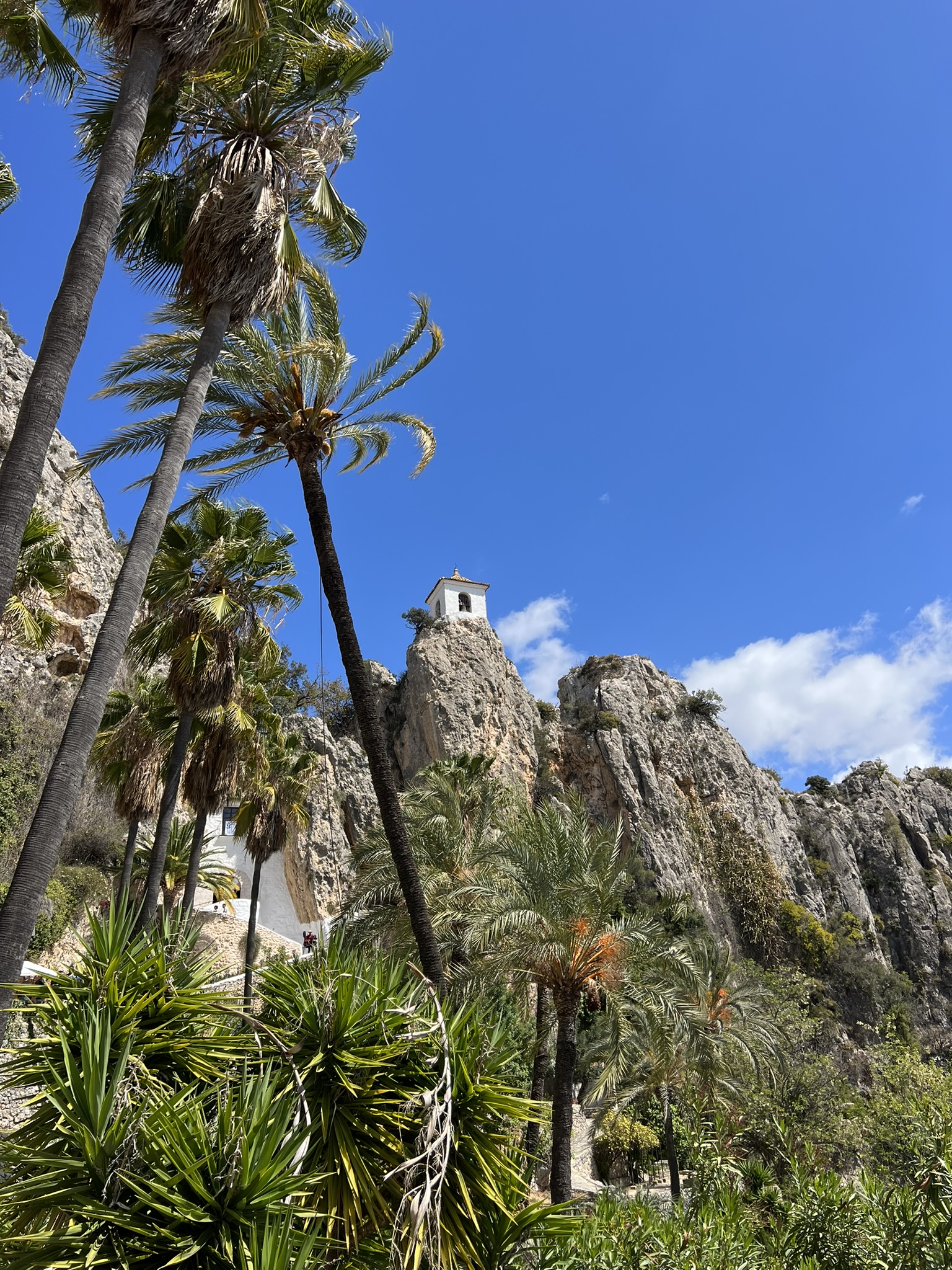 Der Glockenturm von Guadalest auf seiner Felsspitze, gesehen durch hohe Palmen, mit einem kleinen weiß getünchten Gebäude und Stufen am Fuß des Felsens