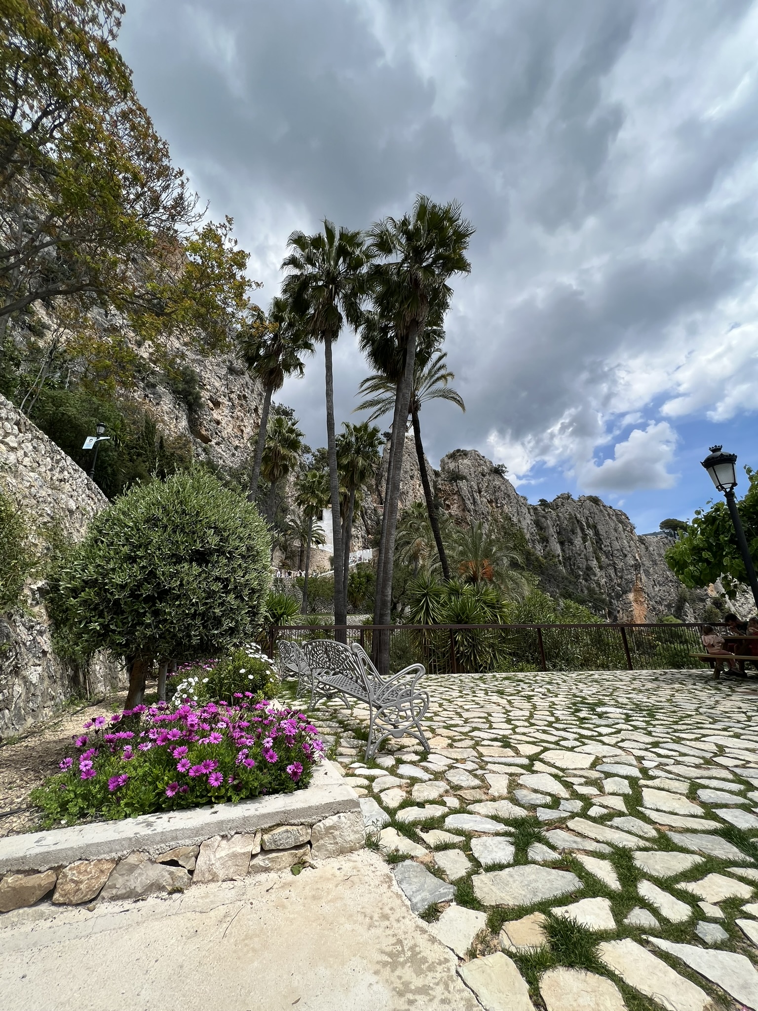 Eine gepflasterte Gartenterrasse in Guadalest mit weißen Metallbänken und violetten Gänseblümchen, mit Blick auf die Berge