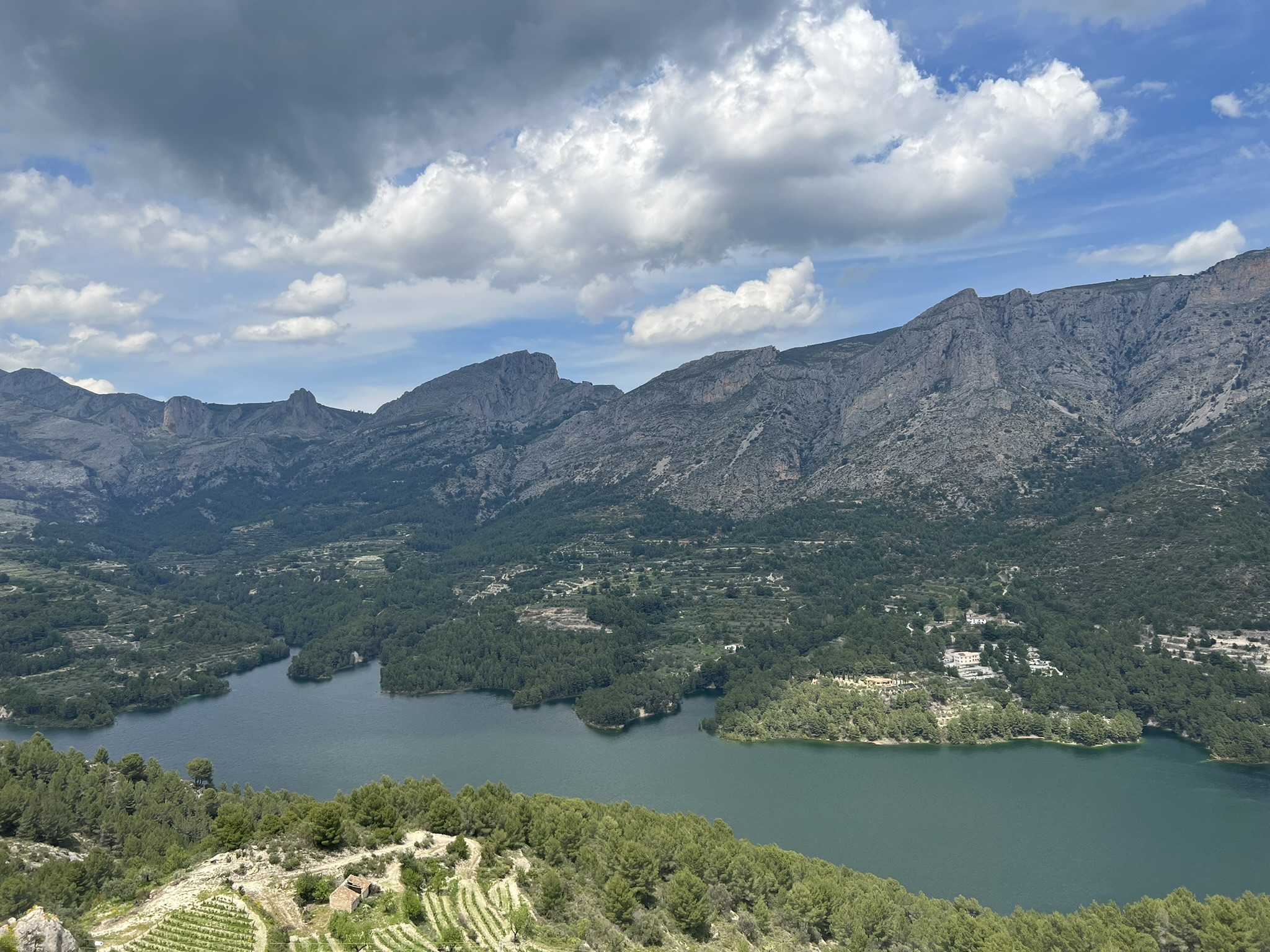 Weiter Blick auf den Stausee Embalse de Guadalest mit bewaldeten Inseln und weißen Dorfhäusern am gegenüberliegenden Ufer