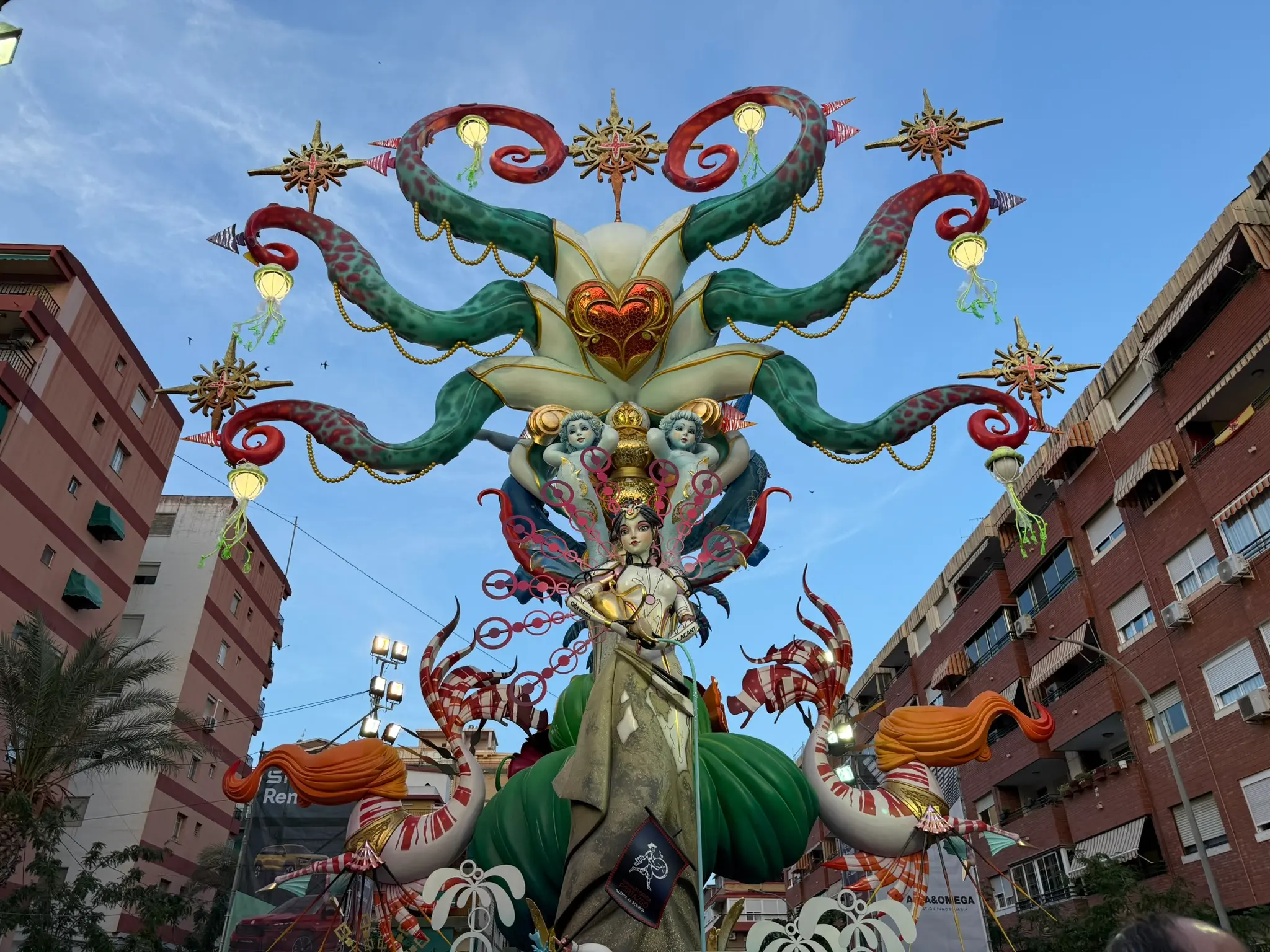 Grand official Hogueras de San Juan monument (Fogueres figure) in Alicante, Spain, under a clear sky — a complex, multi-tiered papier-mâché sculpture surrounded by palm trees
