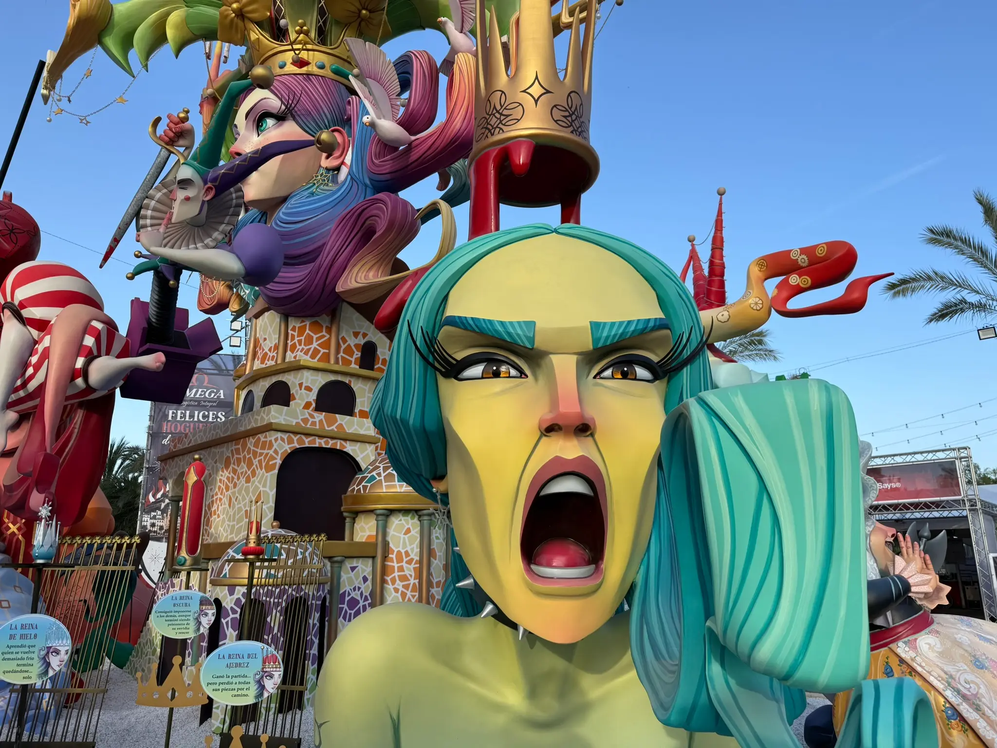 A detailed view of a large, complex Hogueras monument featuring a green-haired figure screaming in the foreground and an elaborate queen character with a golden crown, under a clear blue sky