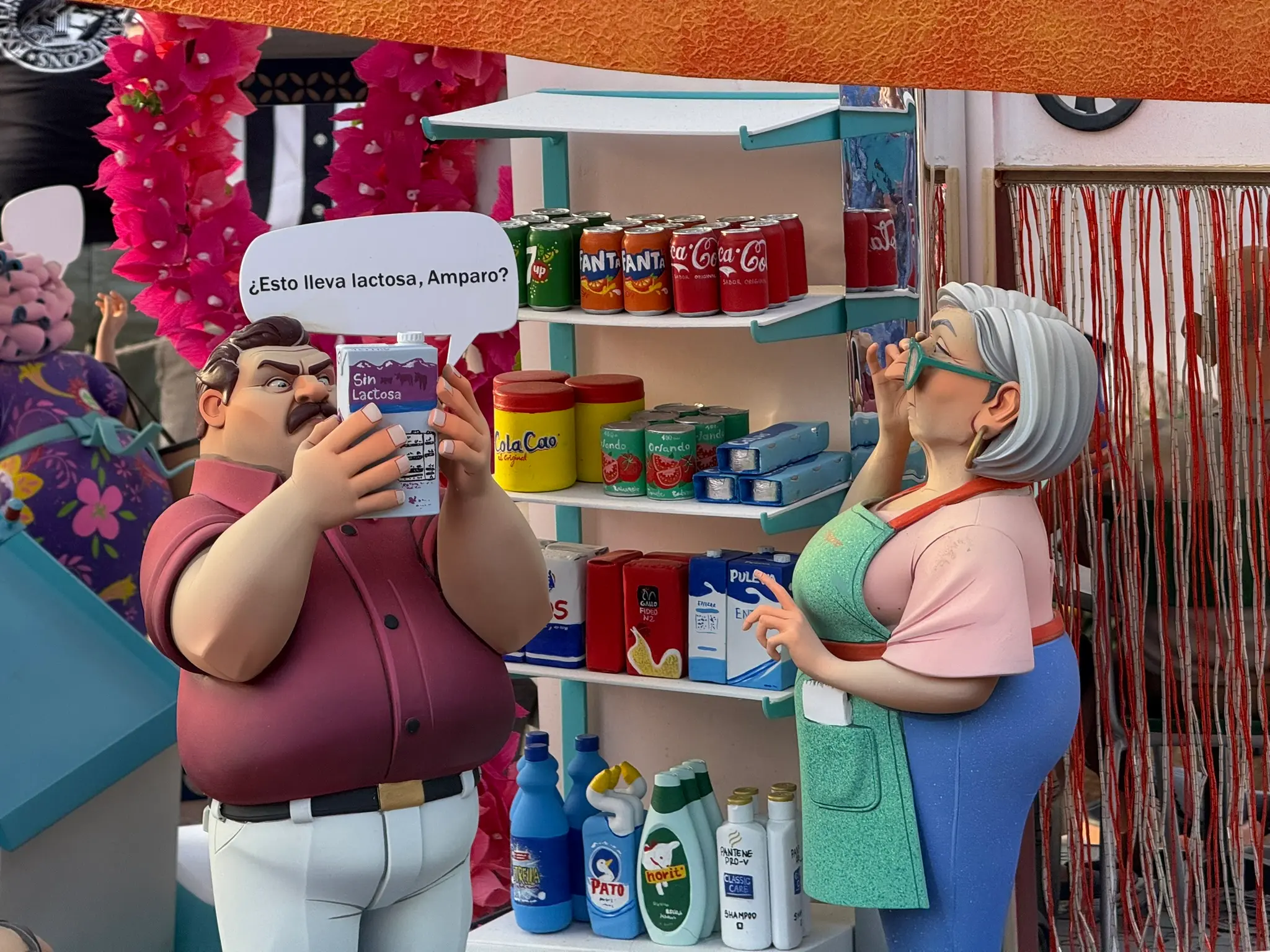 A satirical ninot scene featuring a man holding a 'Sin Lactosa' milk carton speaking to a woman behind a deli counter, with a street sign for 'Carrer de la Sàtira'