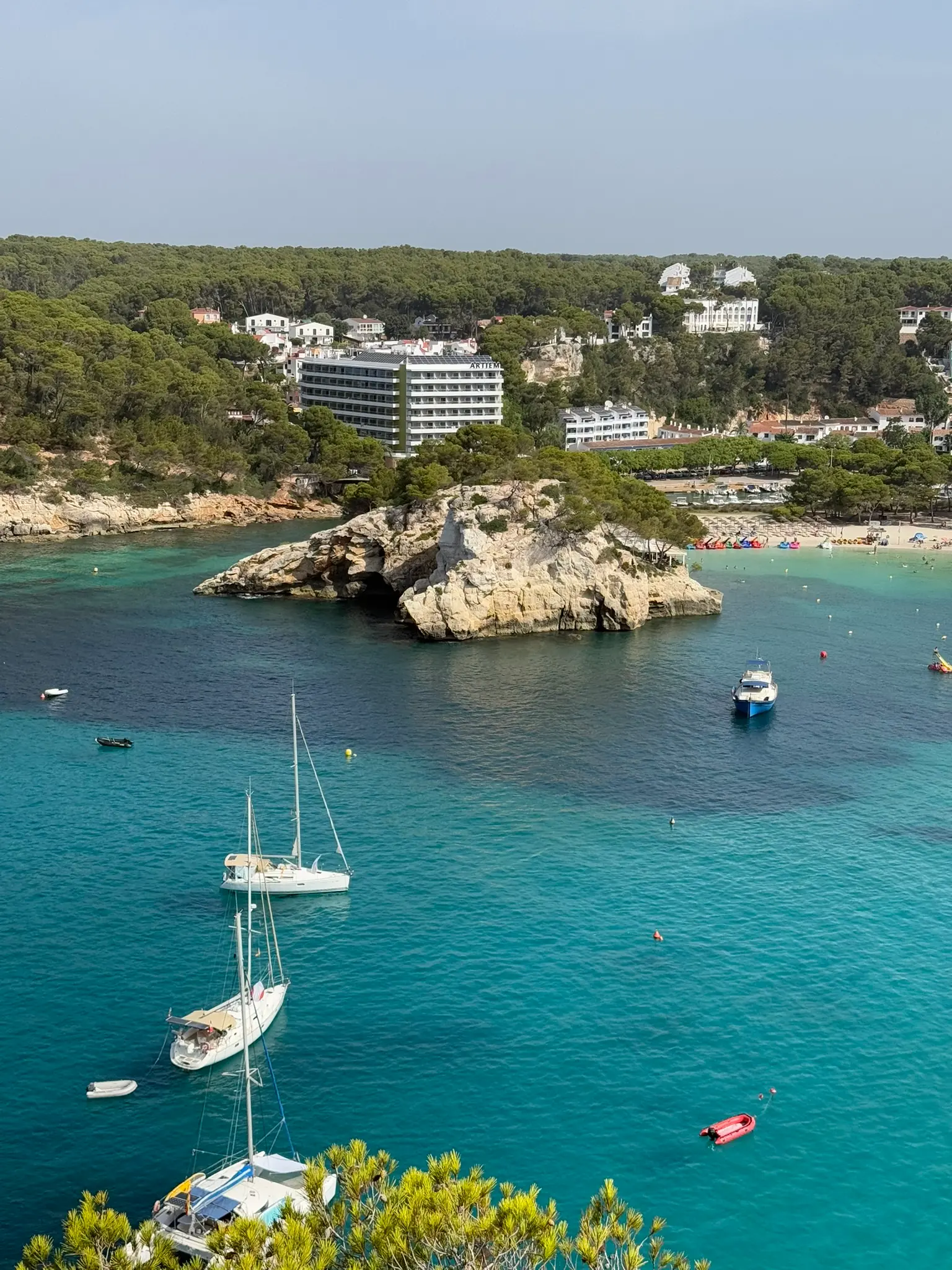 Een brede baai op Menorca met verankerde zeilboten, een wit zandstrand en een hotel op de landtong op de achtergrond