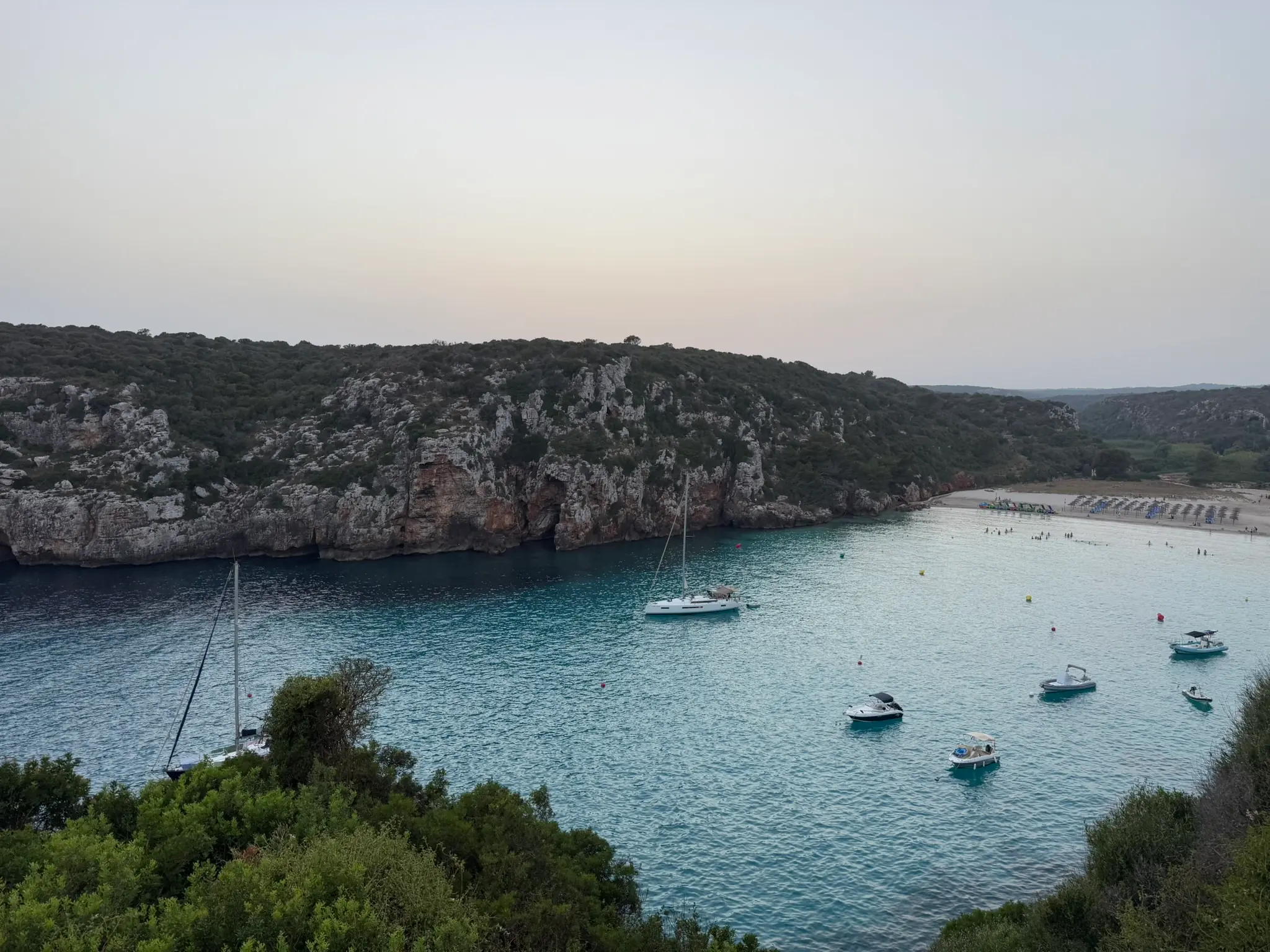 Een baai op Menorca bij schemering, zeilboten voor anker onder pijnboombegroeide kliffen