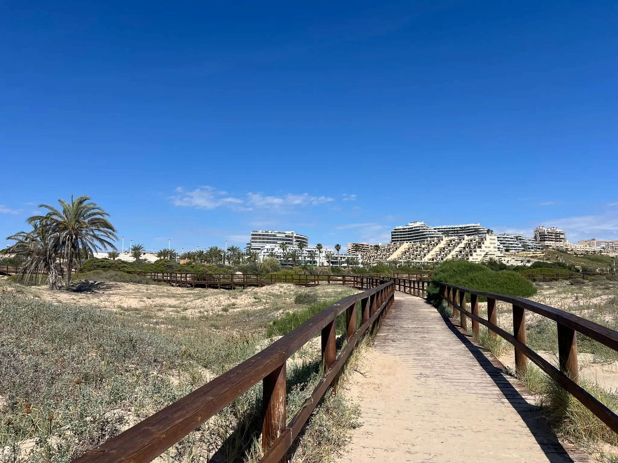 A scenic wooden boardwalk leading through the sand dunes toward the pristine Platja del Carabassí beach, with a clear blue sky in the background.