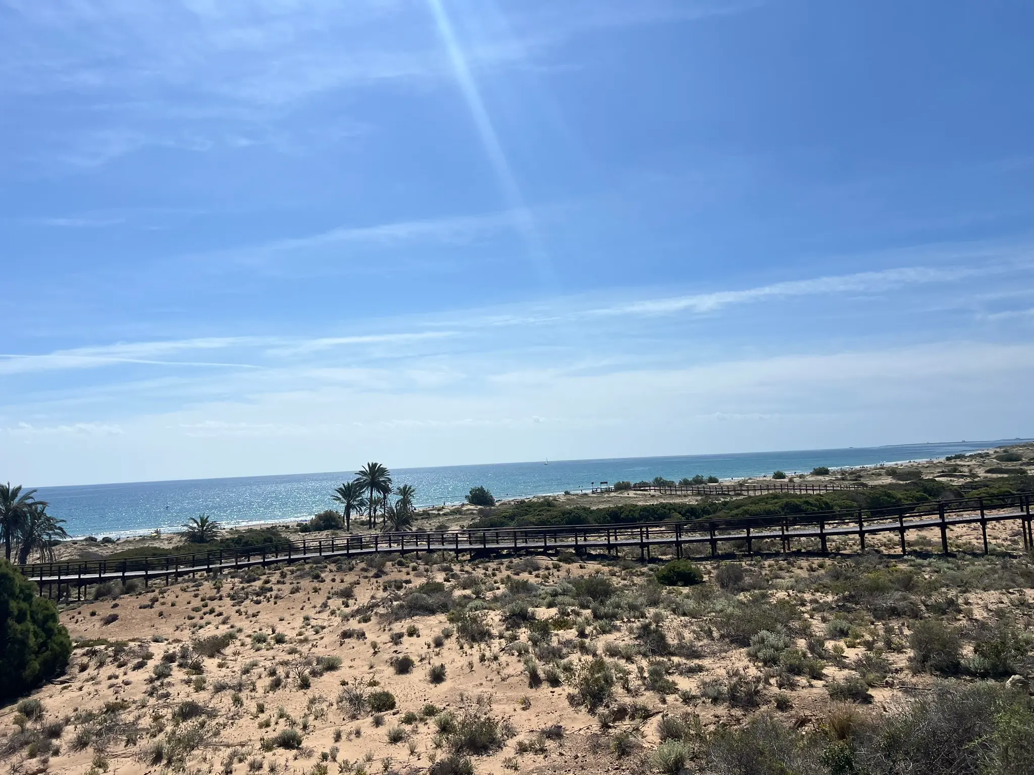 A high-angle, wide panoramic photograph capturing the vast expanse of golden sands and the sparkling Mediterranean Sea at Platja del Carabassí beach in Alicante, with the dune ecosystem and a wooden walkway on the left.