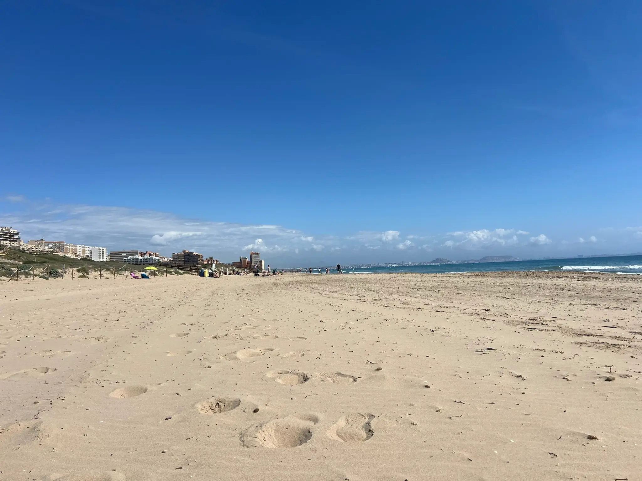 A stunning, low-angle perspective of the expansive golden sands at Platja del Carabassí beach in Alicante, Spain, leading toward the gentle, turquoise waves of the Mediterranean Sea.