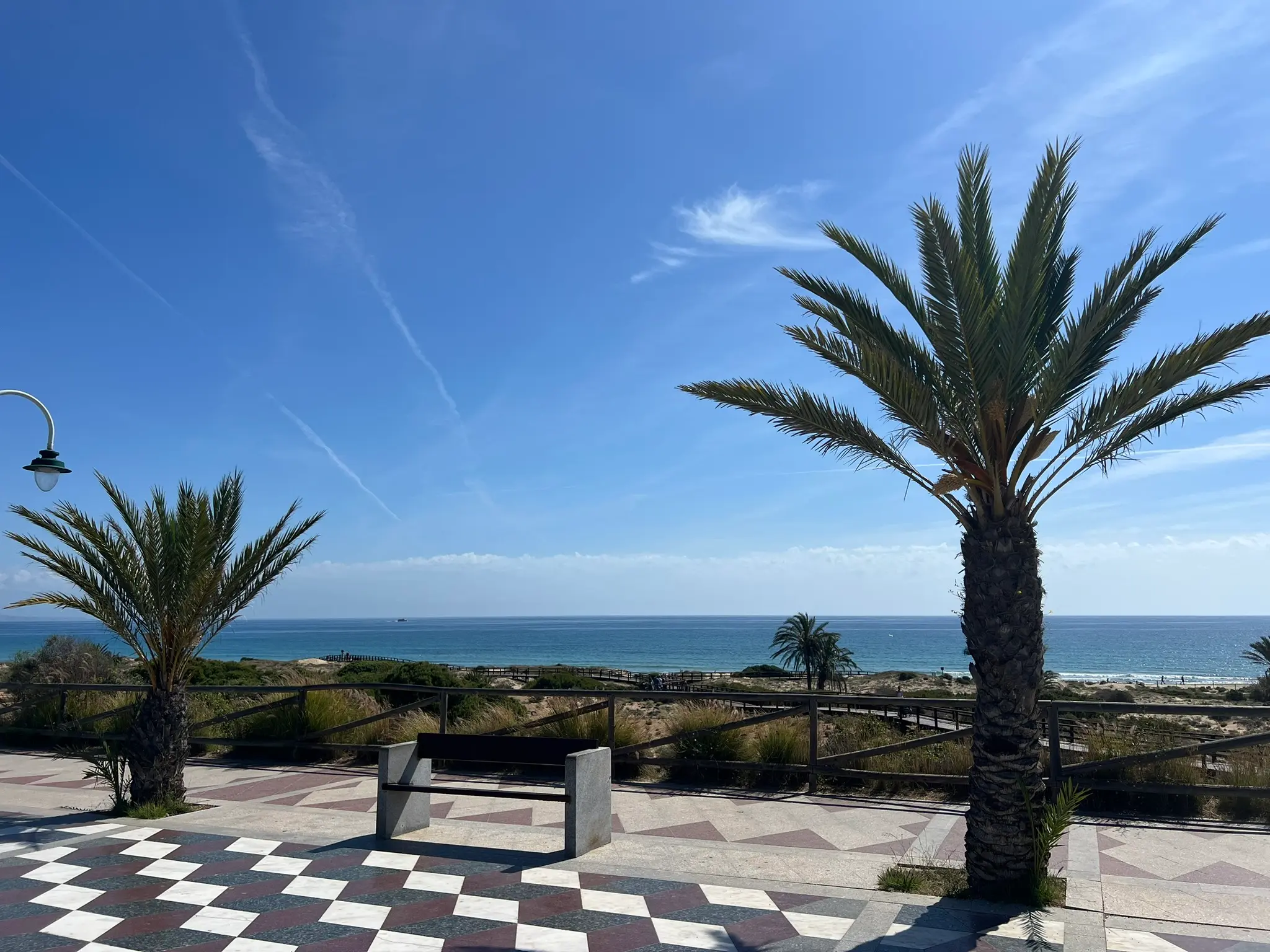 A breathtaking view from the scenic promenade at Platja del Carabassí beach in Alicante, Spain, on a bright, sunny day, with palm trees framing the checkered tiled path.