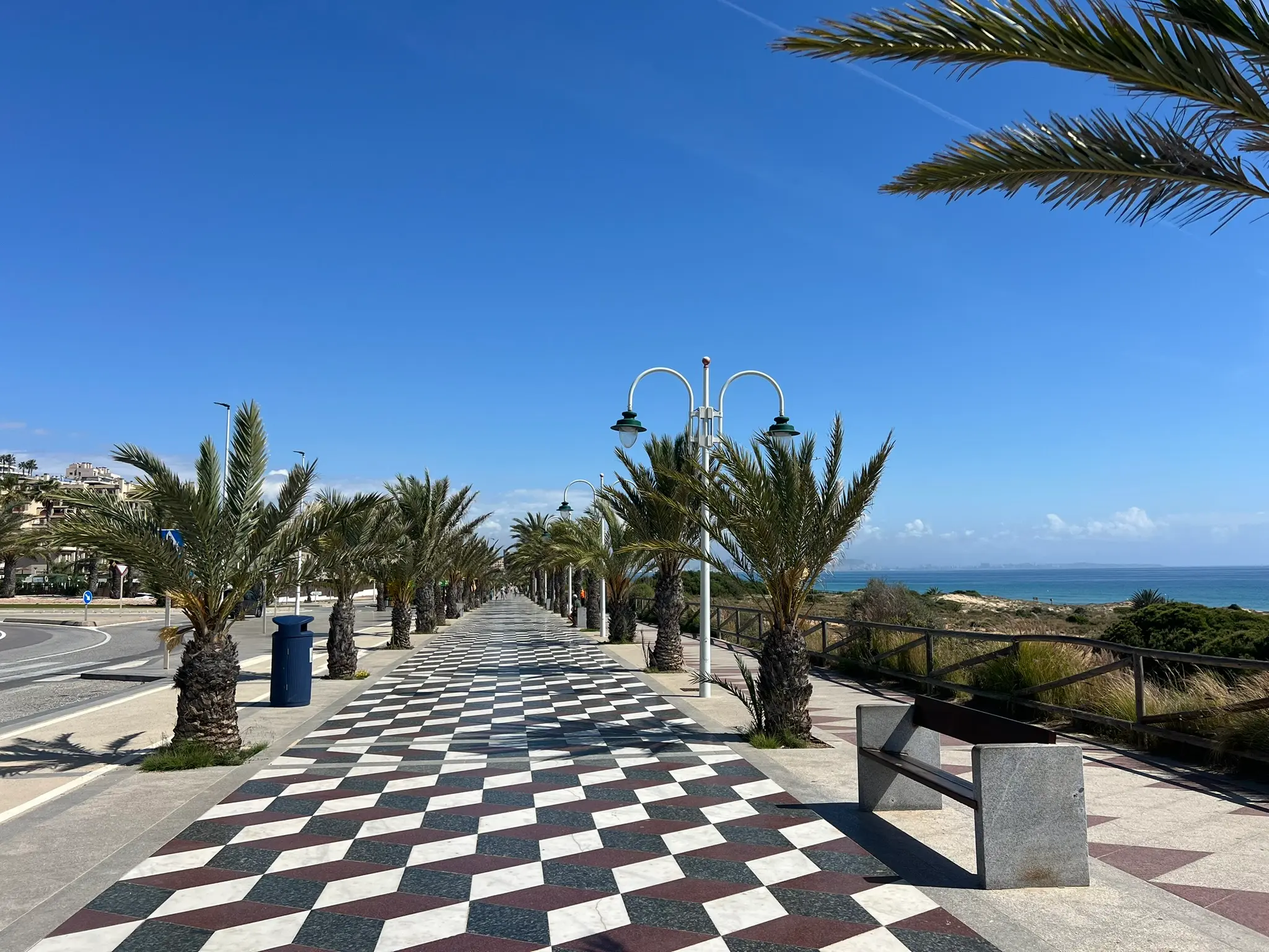 A high-quality, sunlit perspective of the iconic coastal promenade at Platja del Carabassí beach in Alicante, featuring a black-and-white checkered tile design and palm trees.