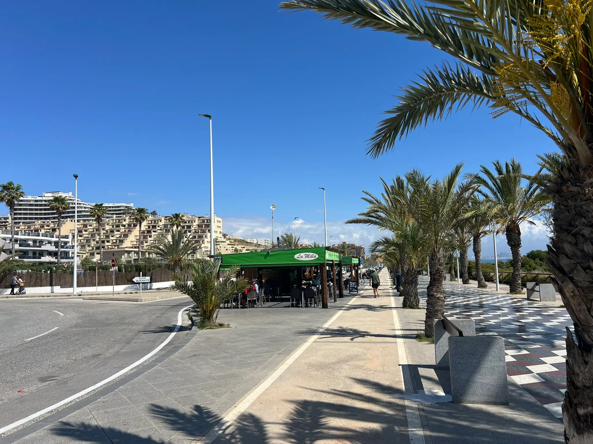A lively, sun-drenched promenade at Platja del Carabassí beach in Alicante, showcasing the iconic black-and-white checkered tile pattern, palm trees, and the La Milla beach cafe.