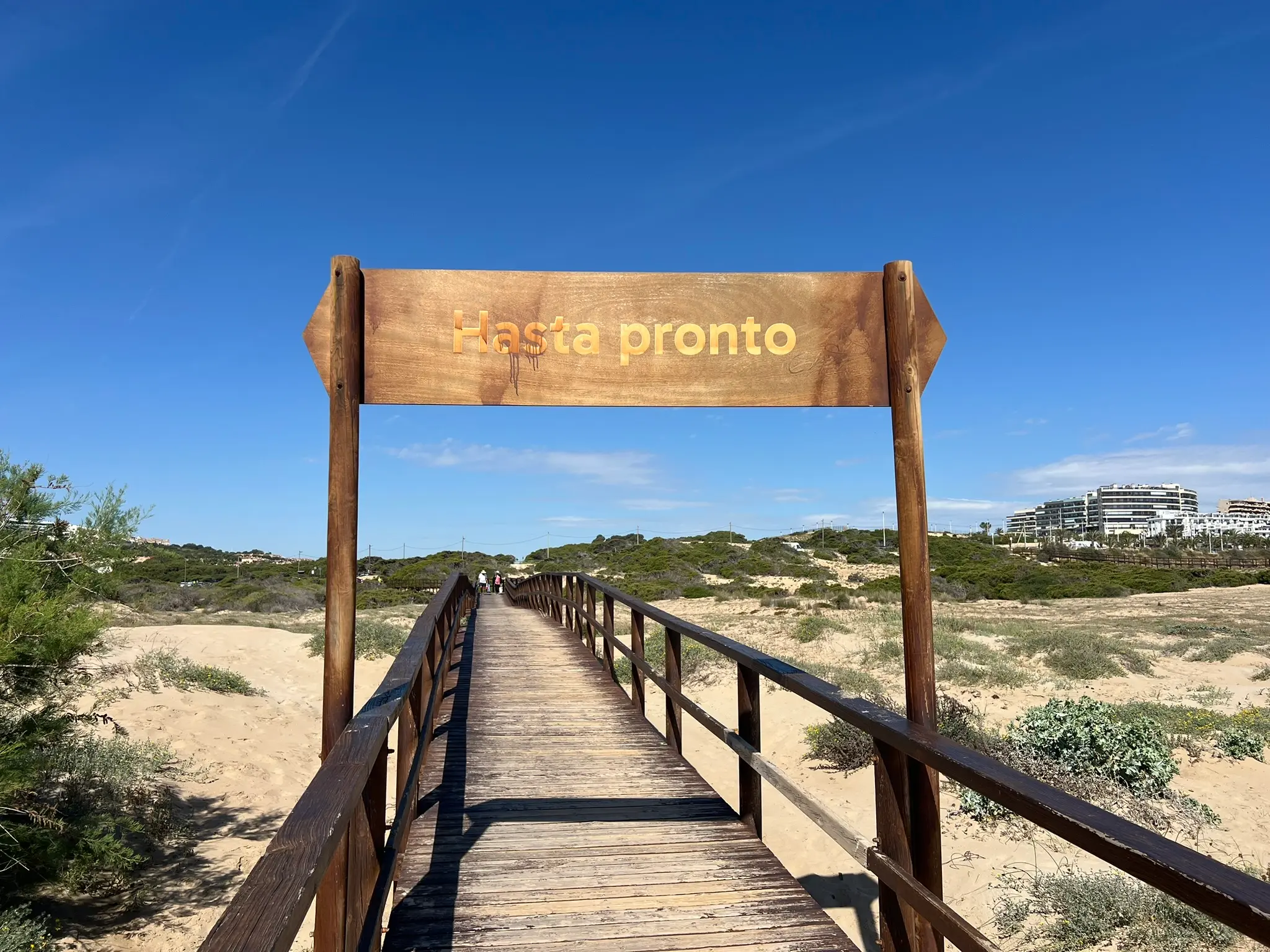 A scenic wooden boardwalk leading away from the Platja del Carabassí beach in Alicante, marked by a rustic wooden sign that reads "Hasta pronto" (See you soon).