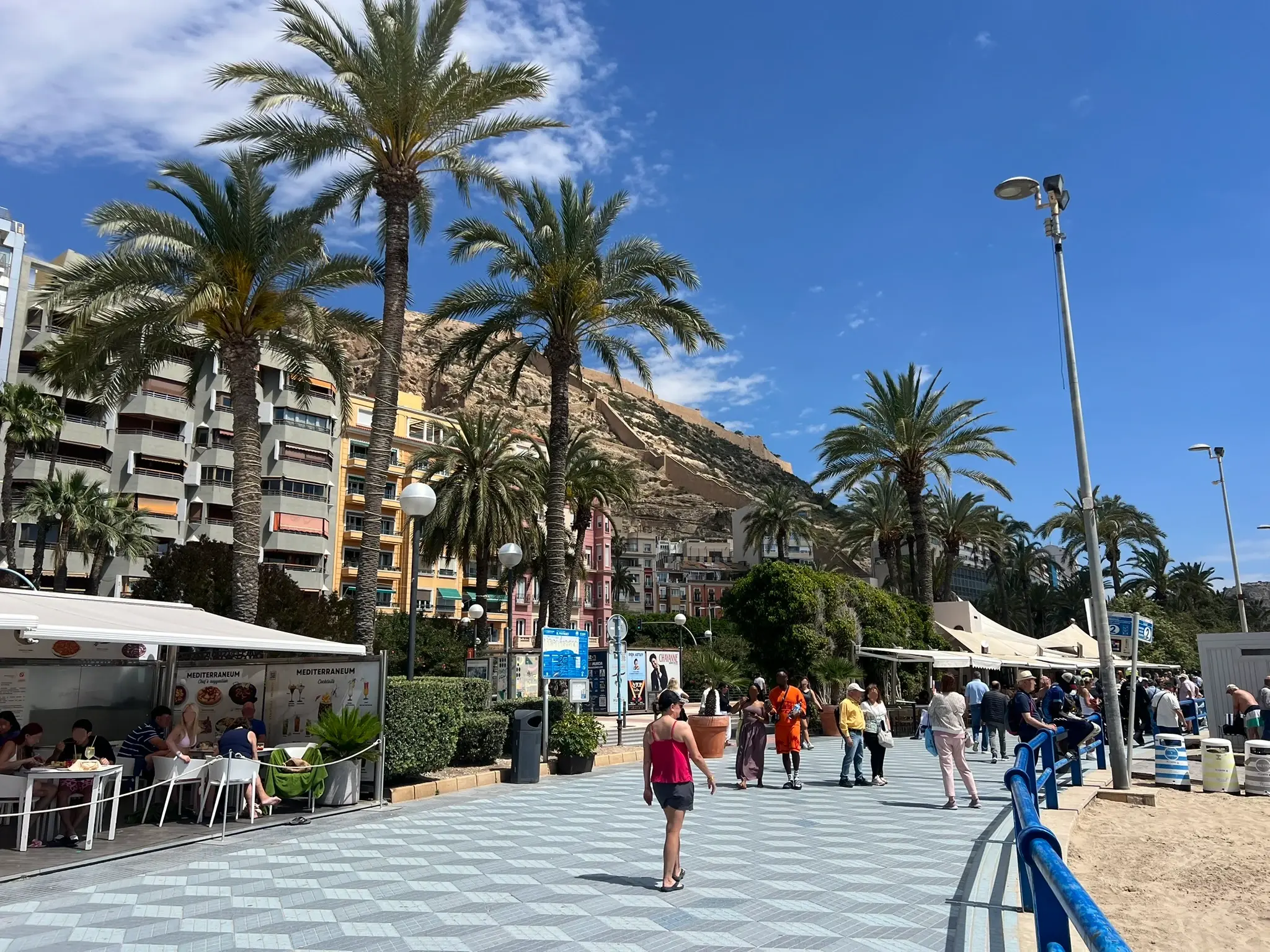 Playa del Postiguet Alicante — busy beachfront promenade with outdoor restaurants, palm trees and Santa Barbara Castle towering above Alicante city centre