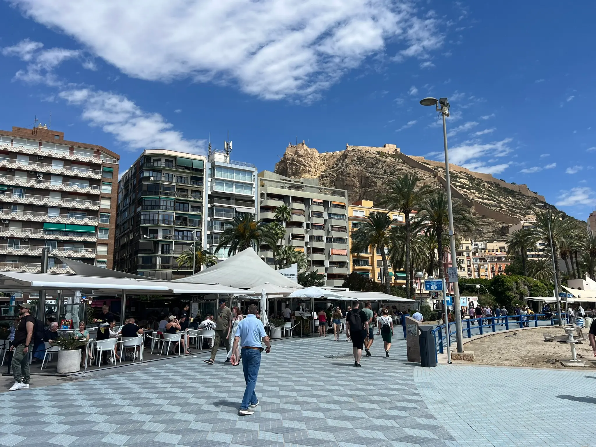 Playa del Postiguet Alicante — busy beachfront promenade with Mediterraneum restaurant, tall palm trees and Santa Barbara Castle rising above Alicante city centre
