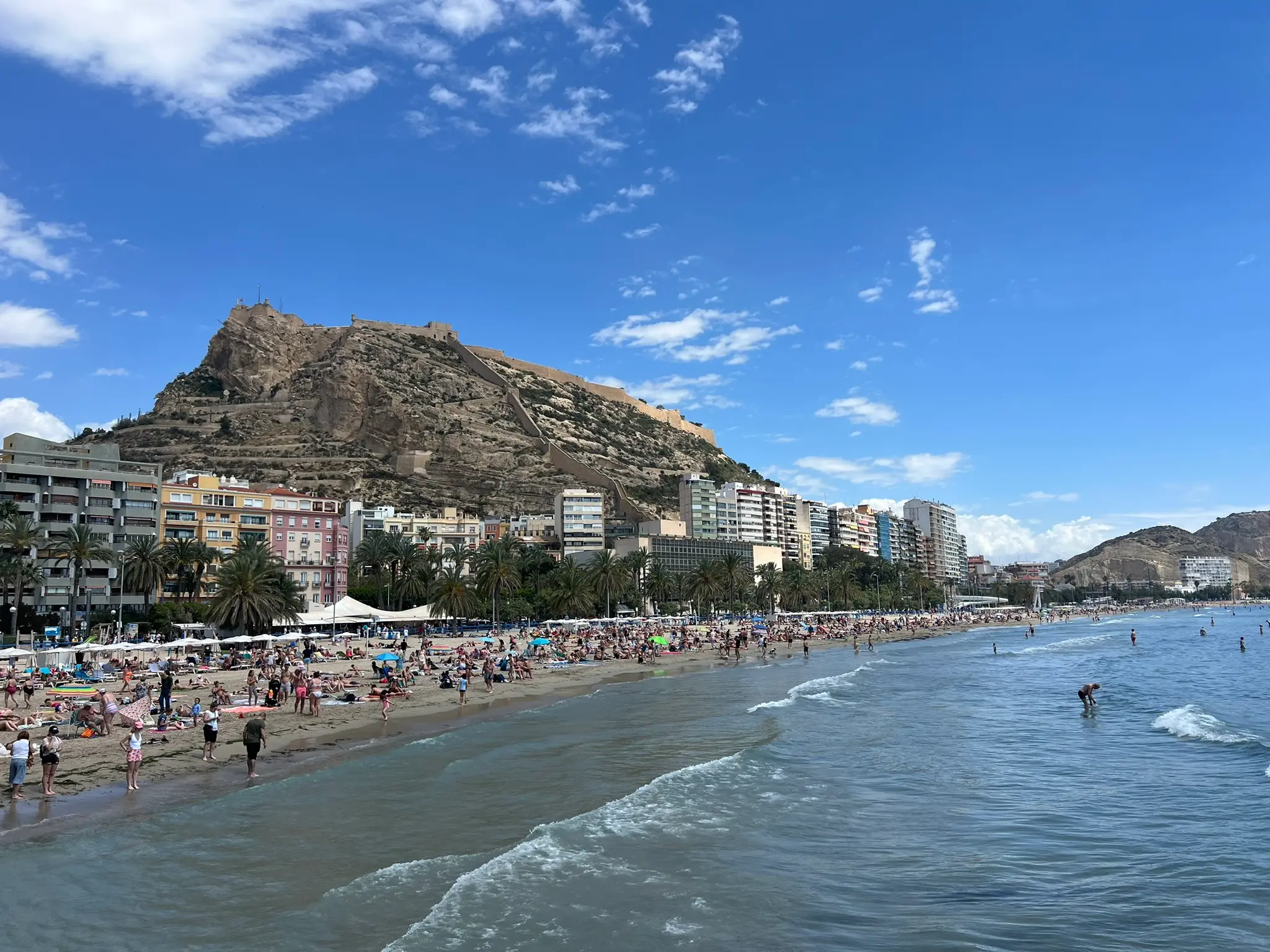 Playa del Postiguet Alicante — iconic view of the crowded city beach with Santa Barbara Castle dramatically rising above Alicante city skyline and Mediterranean waves