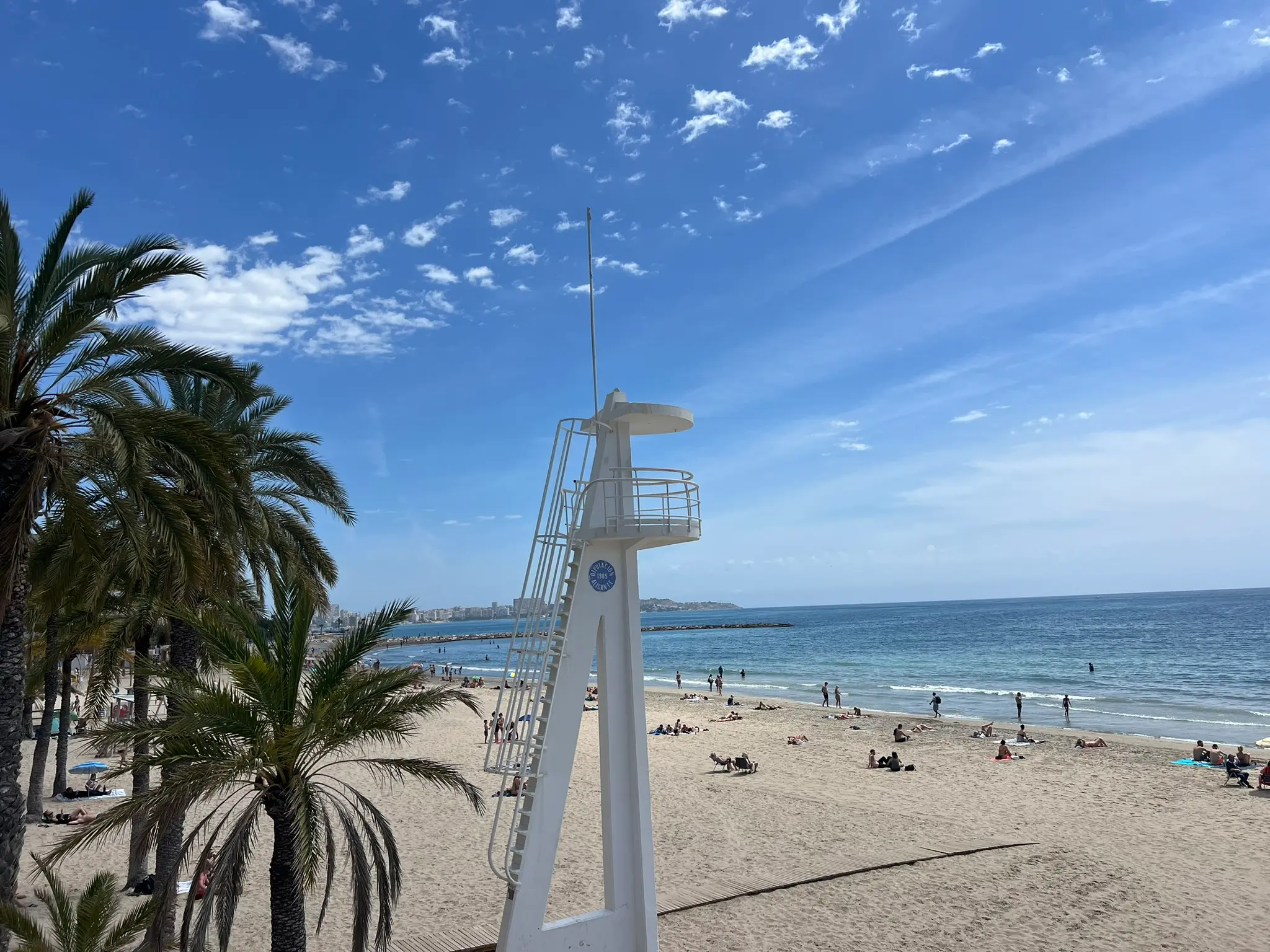 Playa del Postiguet Alicante — lifeguard tower with palm trees and busy city beach full of sunbathers and swimmers, Blue Flag beach in Alicante city centre