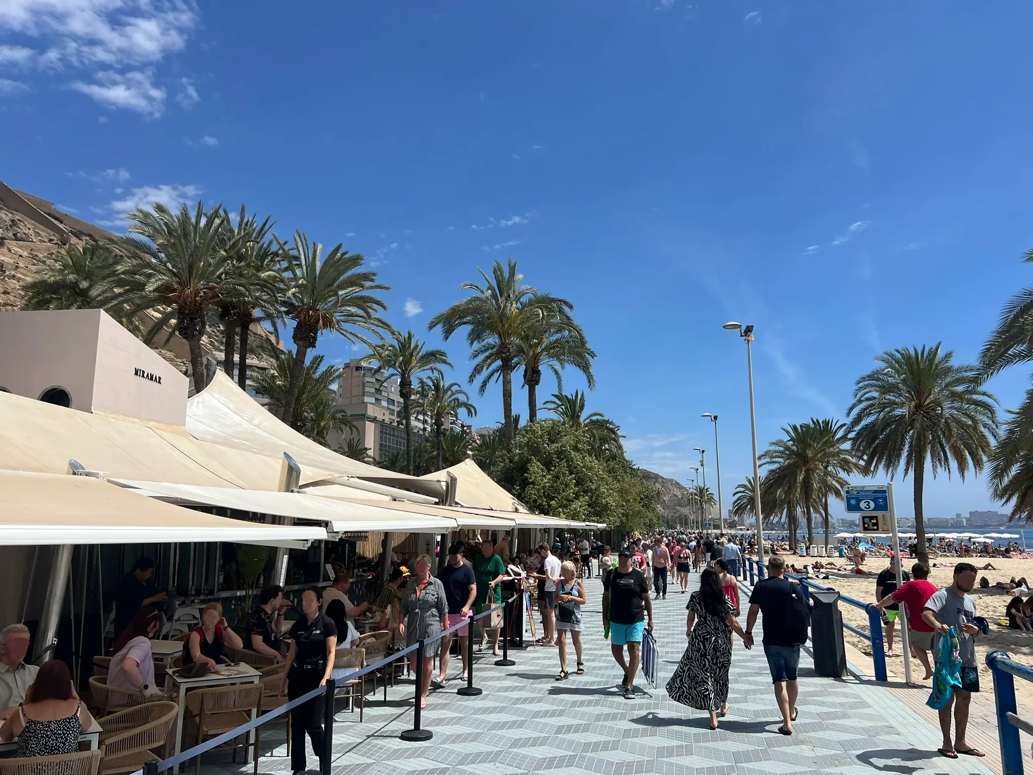 Playa del Postiguet Alicante — busy beachfront promenade with Miramar restaurant, tall palm trees, sunbathers and Santa Barbara Castle in background