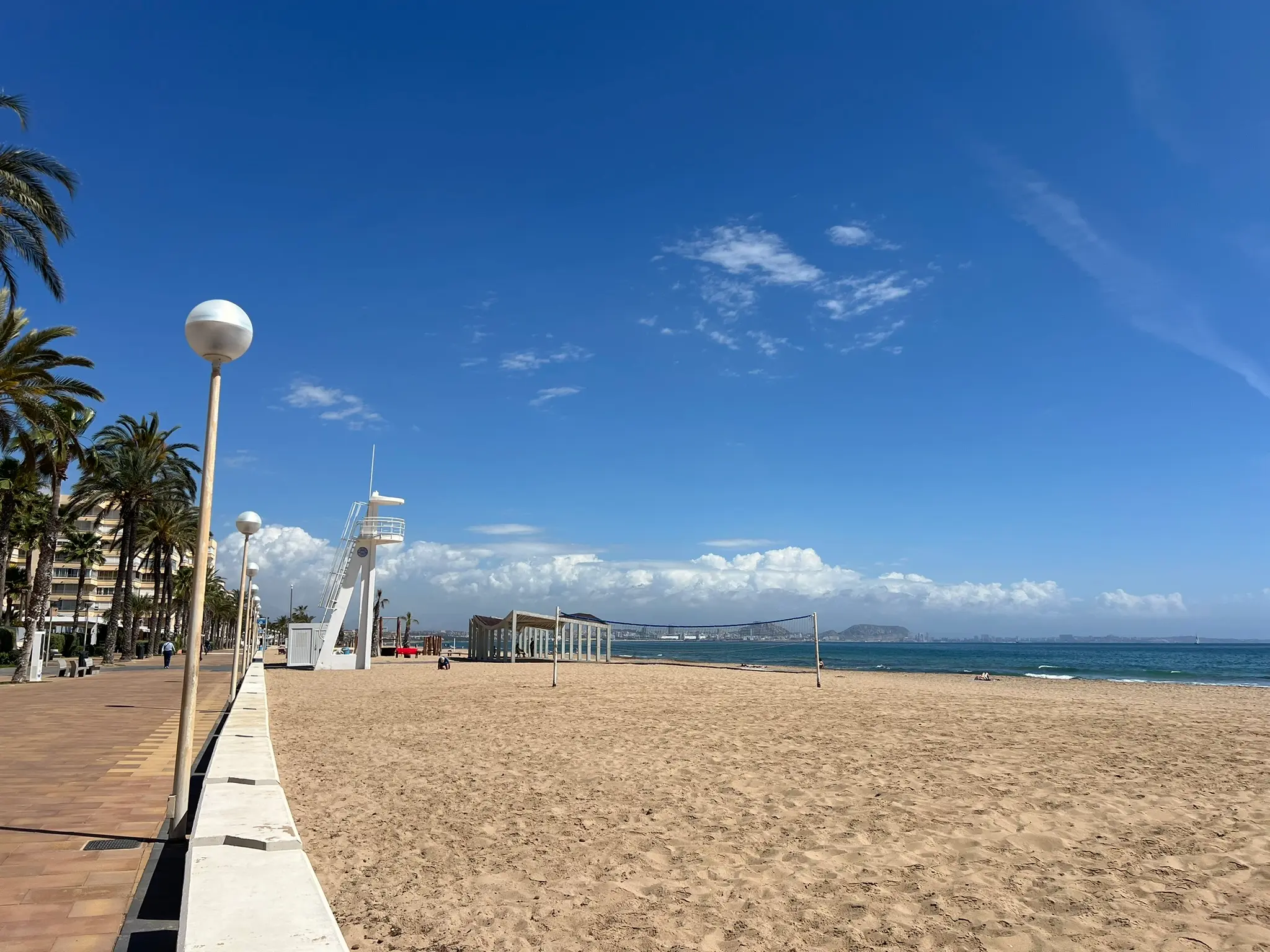 Playa del Saladar Urbanova Alicante — beach volleyball net, lifeguard tower and palm-lined promenade with Alicante skyline in background