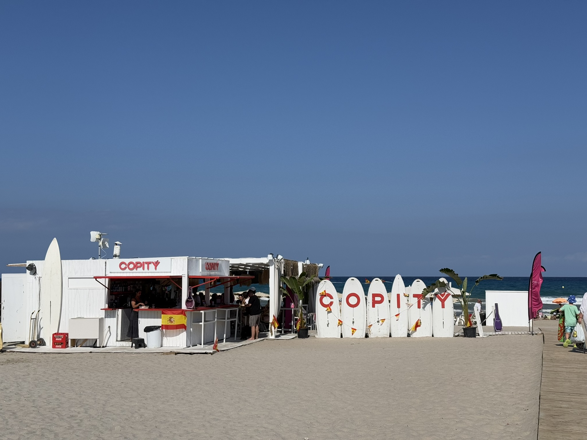 Die Strandbar Copity im Sand der Playa de San Juan, Alicante — ein weißer, mit Surfbrettern verkleideter Kiosk mit spanischer Flagge und Palmenpflanzen, im Hintergrund das Mittelmeer.