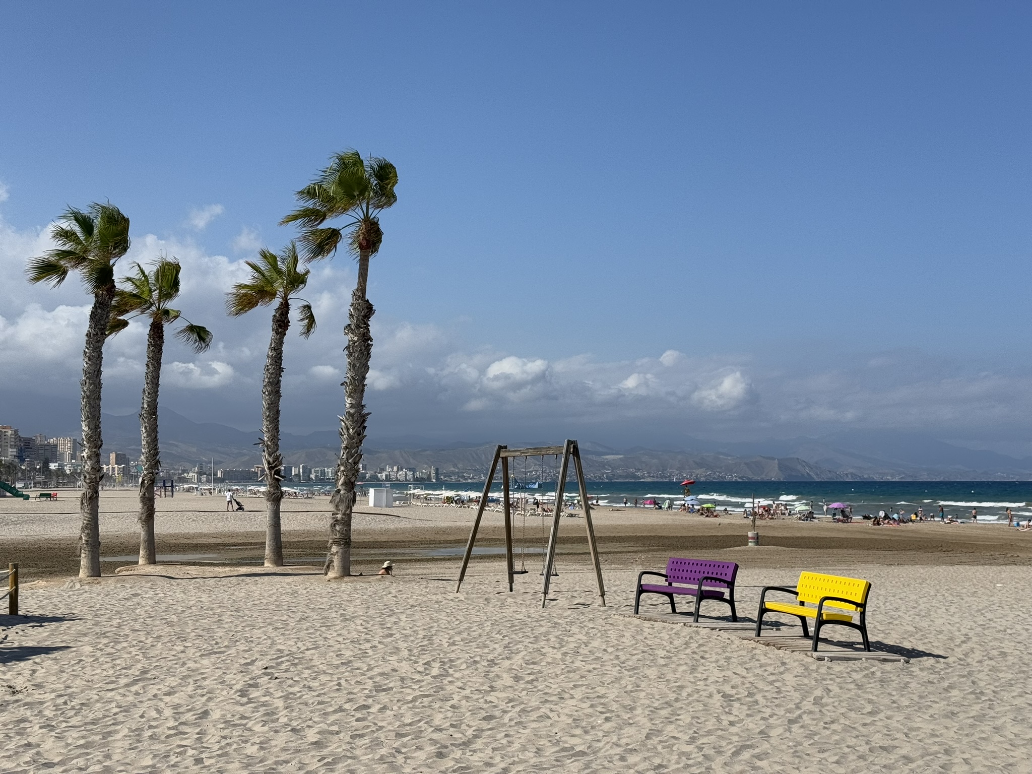 Windbewegte Palmen, eine Schaukel und bunte lila und gelbe Bänke im Sand der Playa de San Juan, Alicante, mit der Küstensilhouette und Bergen in der Ferne.