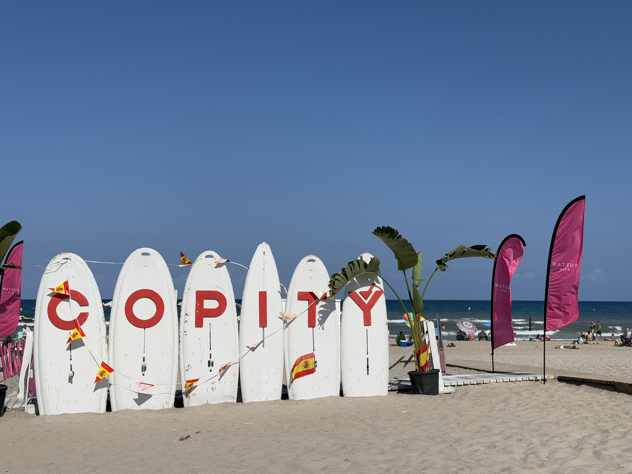 Nahaufnahme des Copity-Strandbar-Schilds an der Playa de San Juan, Alicante — eine Reihe weißer Surfbretter, die in roten Buchstaben COPITY buchstabieren, geschmückt mit spanischer Fahnenkette und Palmenpflanzen.
