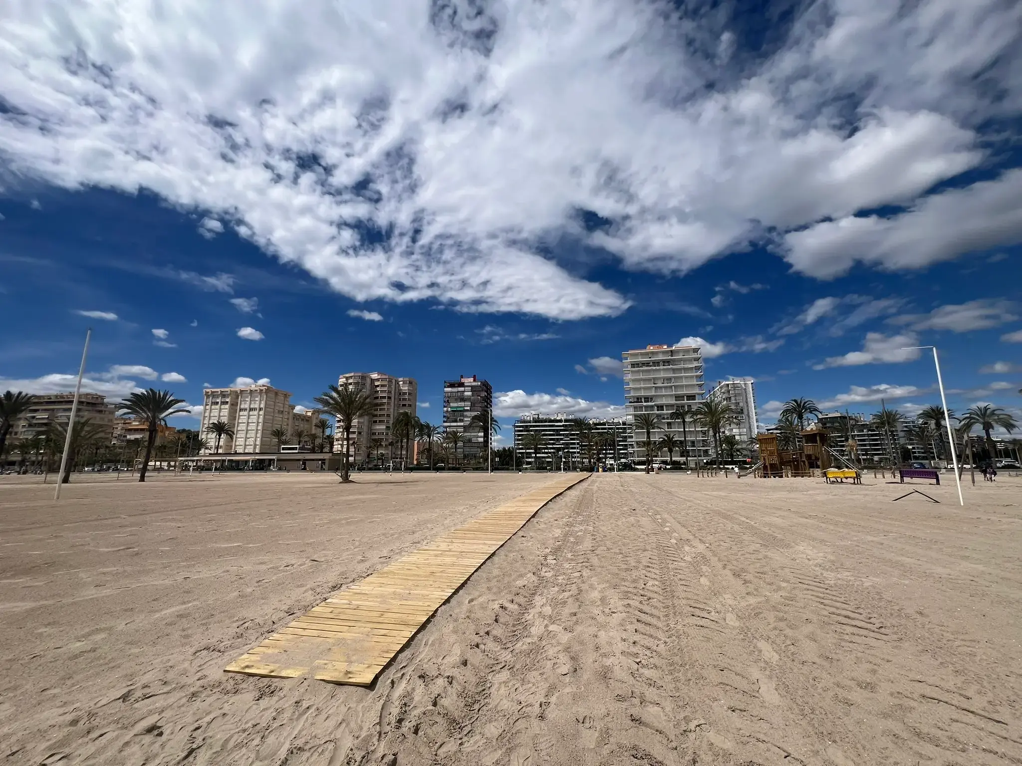 Playa de San Juan Alicante — wooden wheelchair accessible boardwalk on wide sandy beach with apartment buildings, palm trees and children's playground in the background.