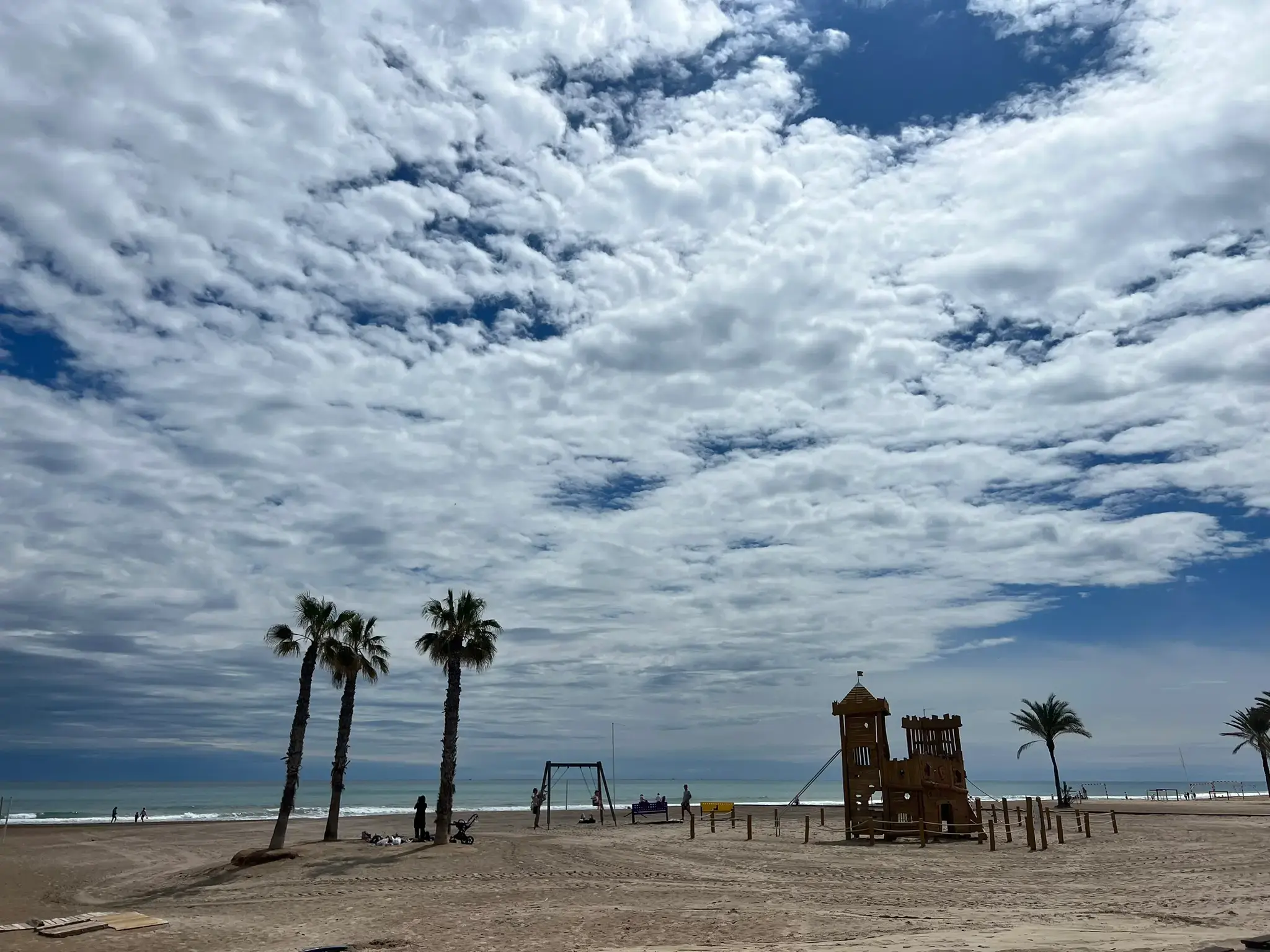 Playa de San Juan Alicante beach — wooden castle playground and swings on wide sandy beach with palm trees and Mediterranean sea under dramatic cloudy sky.