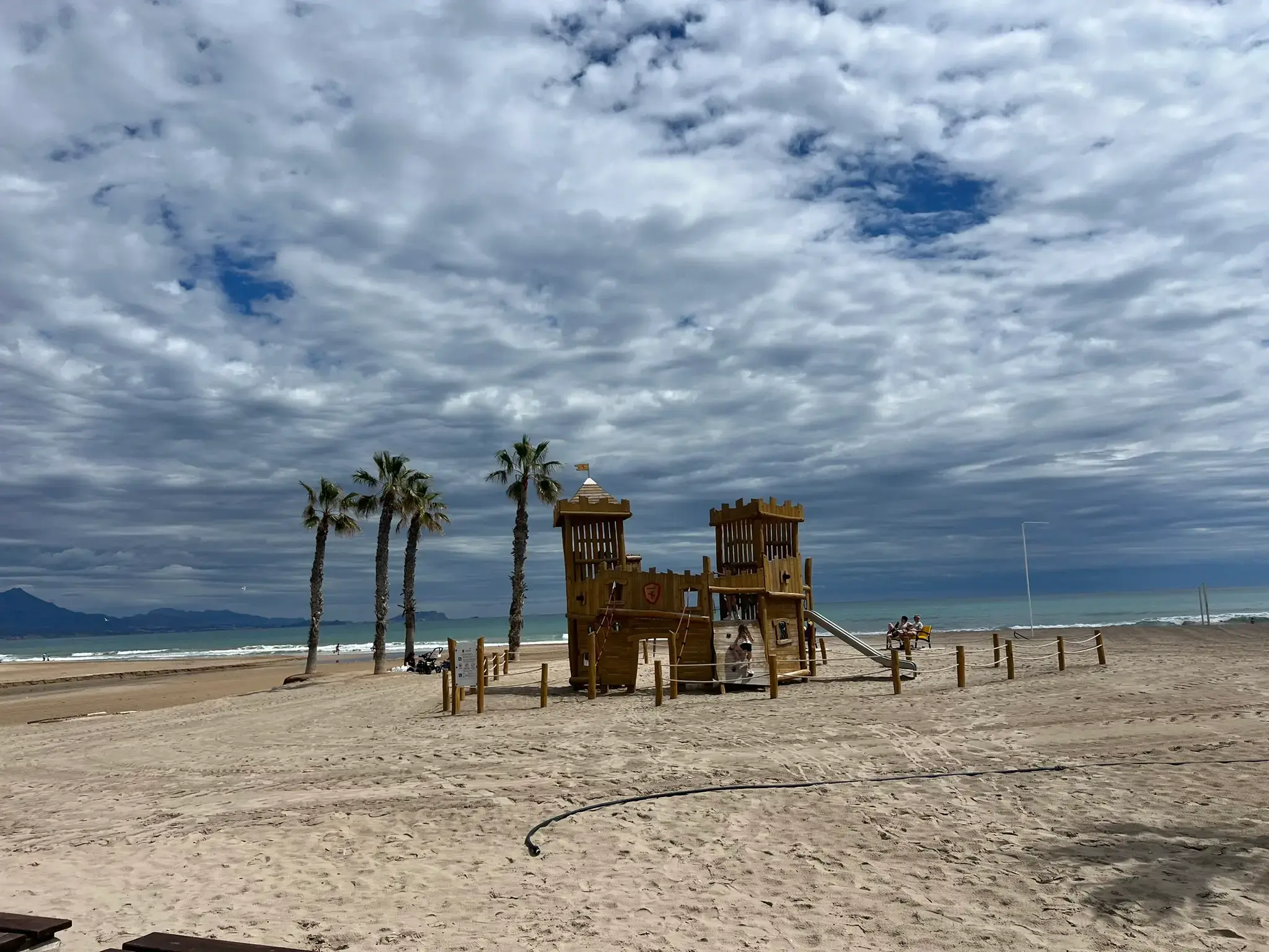 Playa de San Juan Alicante — wooden castle children's playground on the beach with palm trees and Mediterranean sea in the background.