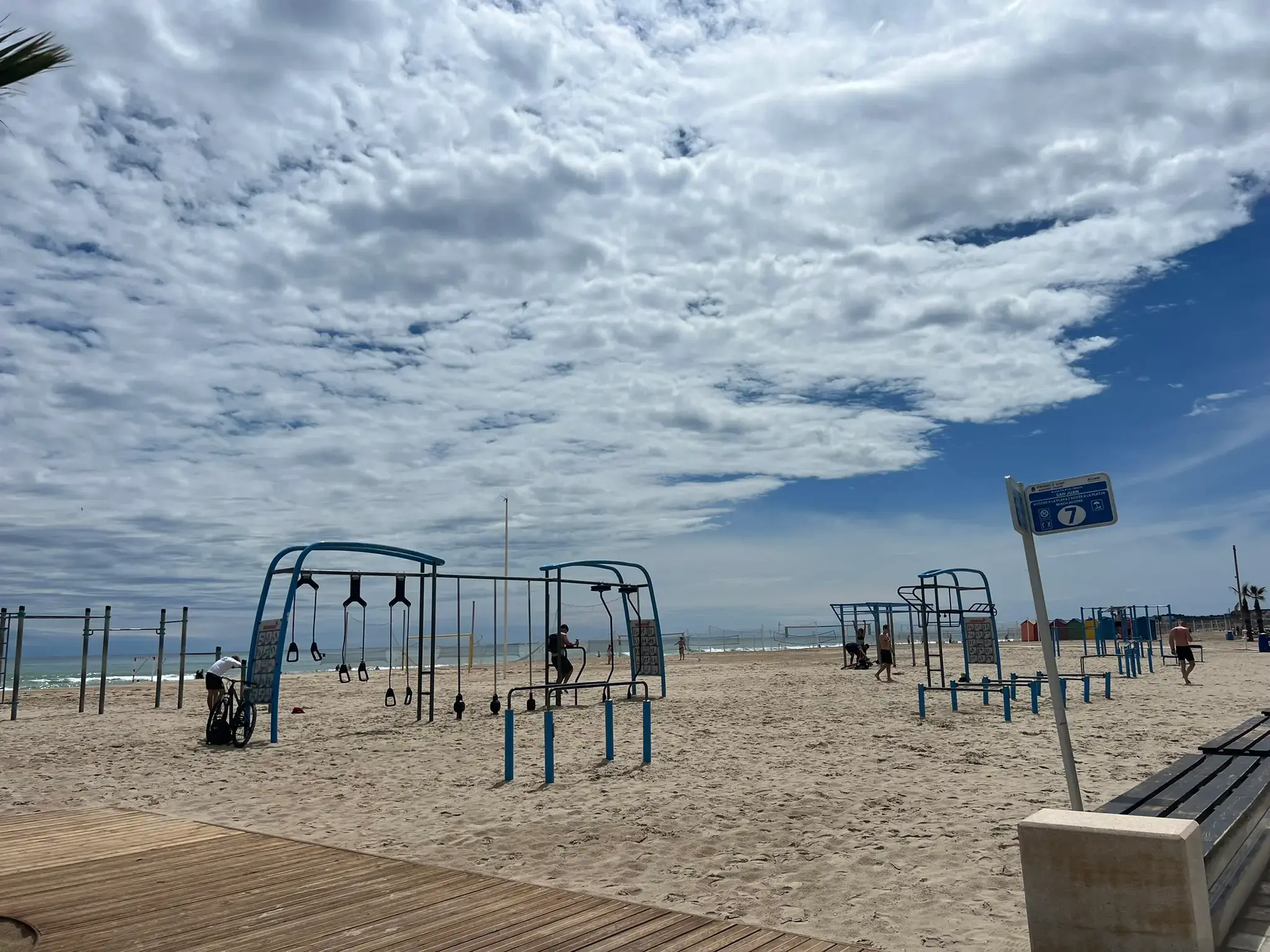 Playa de San Juan Alicante — free outdoor gym and calisthenics equipment on the beach with people exercising, volleyball courts and Mediterranean sea in the background.