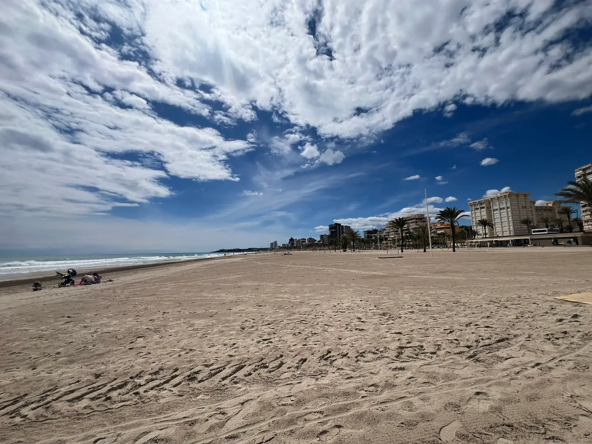Wide sandy expanse of Playa de San Juan in Alicante, Costa Blanca — panoramic shot of the golden shoreline, scattered palm trees, and coastal residential buildings under a dramatic blue sky.