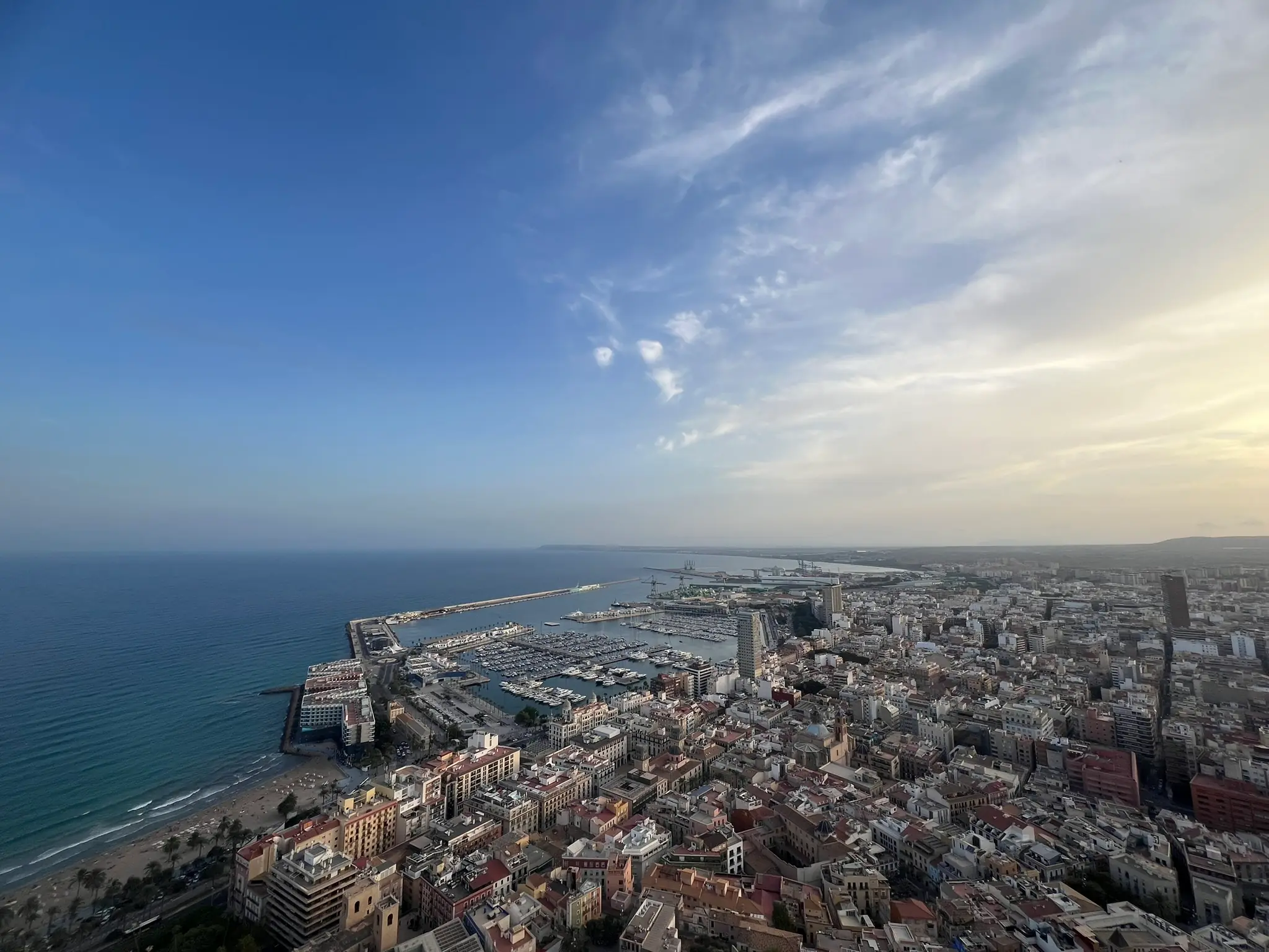 Une vue panoramique aérienne depuis le Castillo de Santa Bárbara sur la ville d'Alicante, le port de plaisance avec ses bateaux, et la côte méditerranéenne au coucher du soleil, avec la plage et la vieille ville historique visibles en contrebas sous un ciel doré du soir.