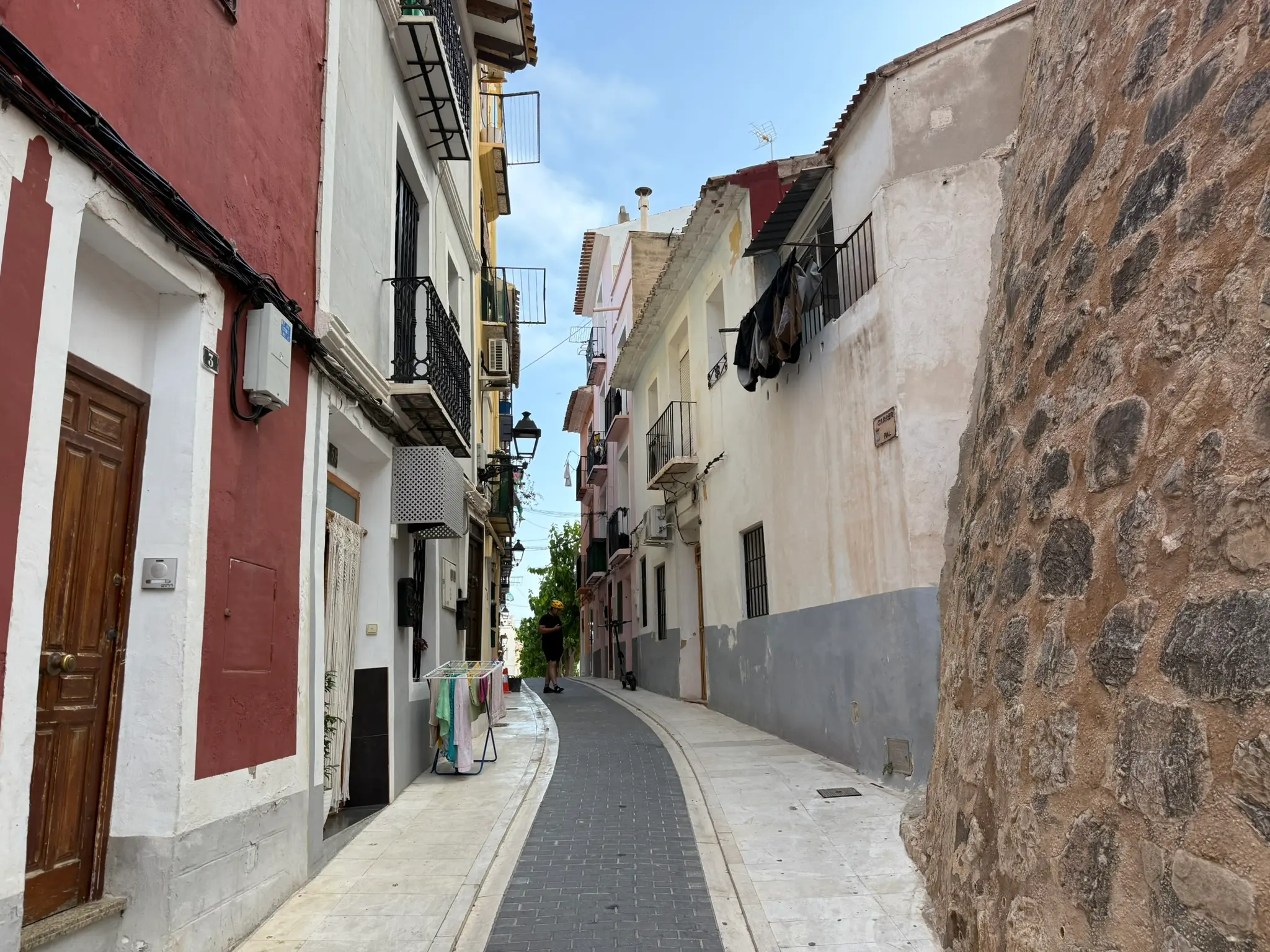 Narrow paved street Carrer del Pal in Villajoyosa — ancient stone defensive wall on right, residential buildings with balconies on left