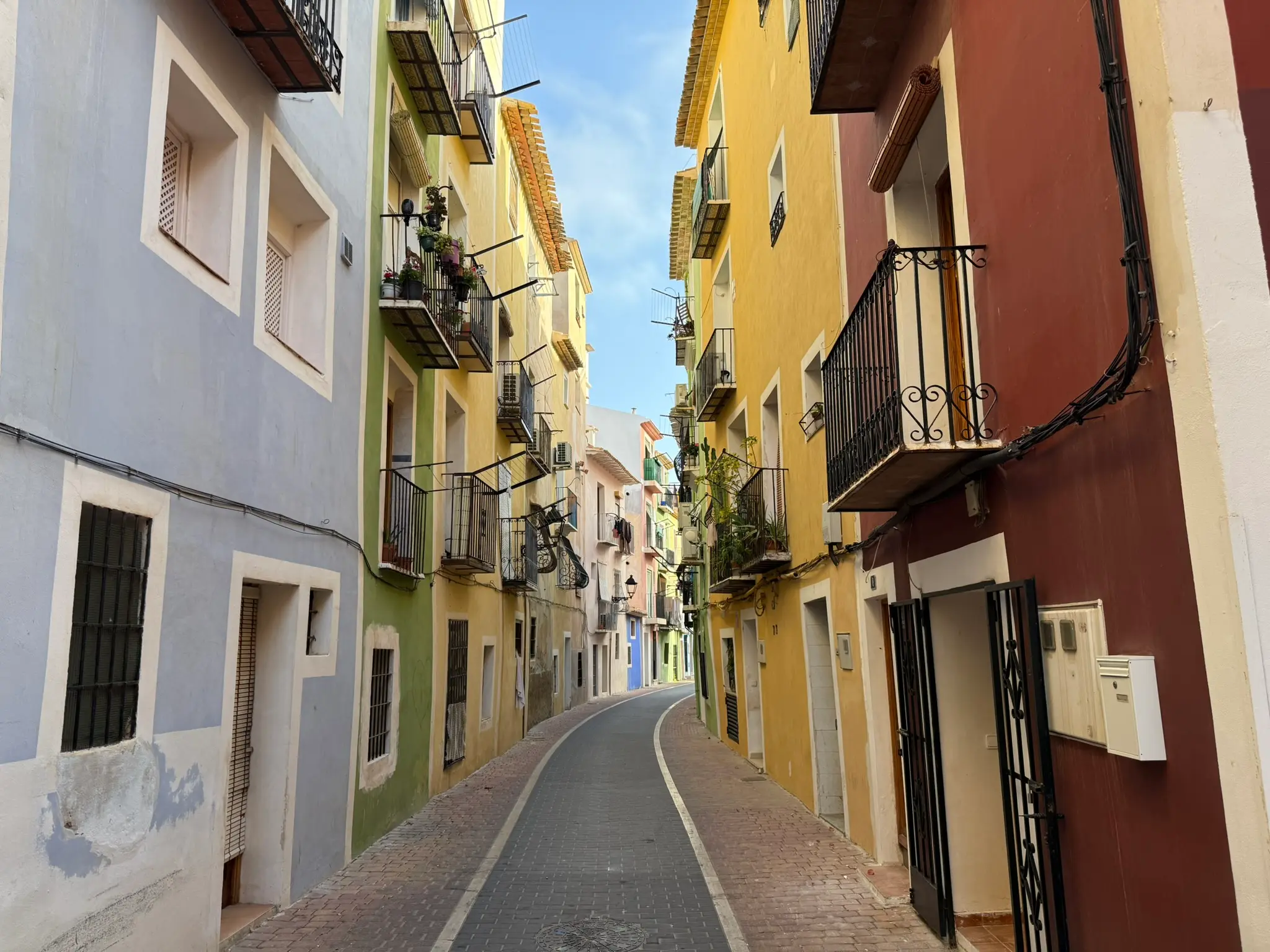 Deep perspective view of narrow curving Carrer Major in Villajoyosa — tall traditional apartment buildings with vibrant blue, green, yellow, and deep red facades with iron balconies