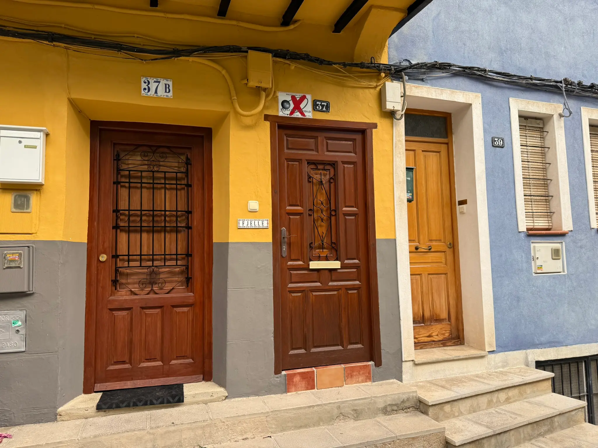 Residential ground floor facades of houses 37, 37B, and 39 in Villajoyosa — bright yellow and blue walls, heavy dark wood doors with iron grates, ceramic family name plaque