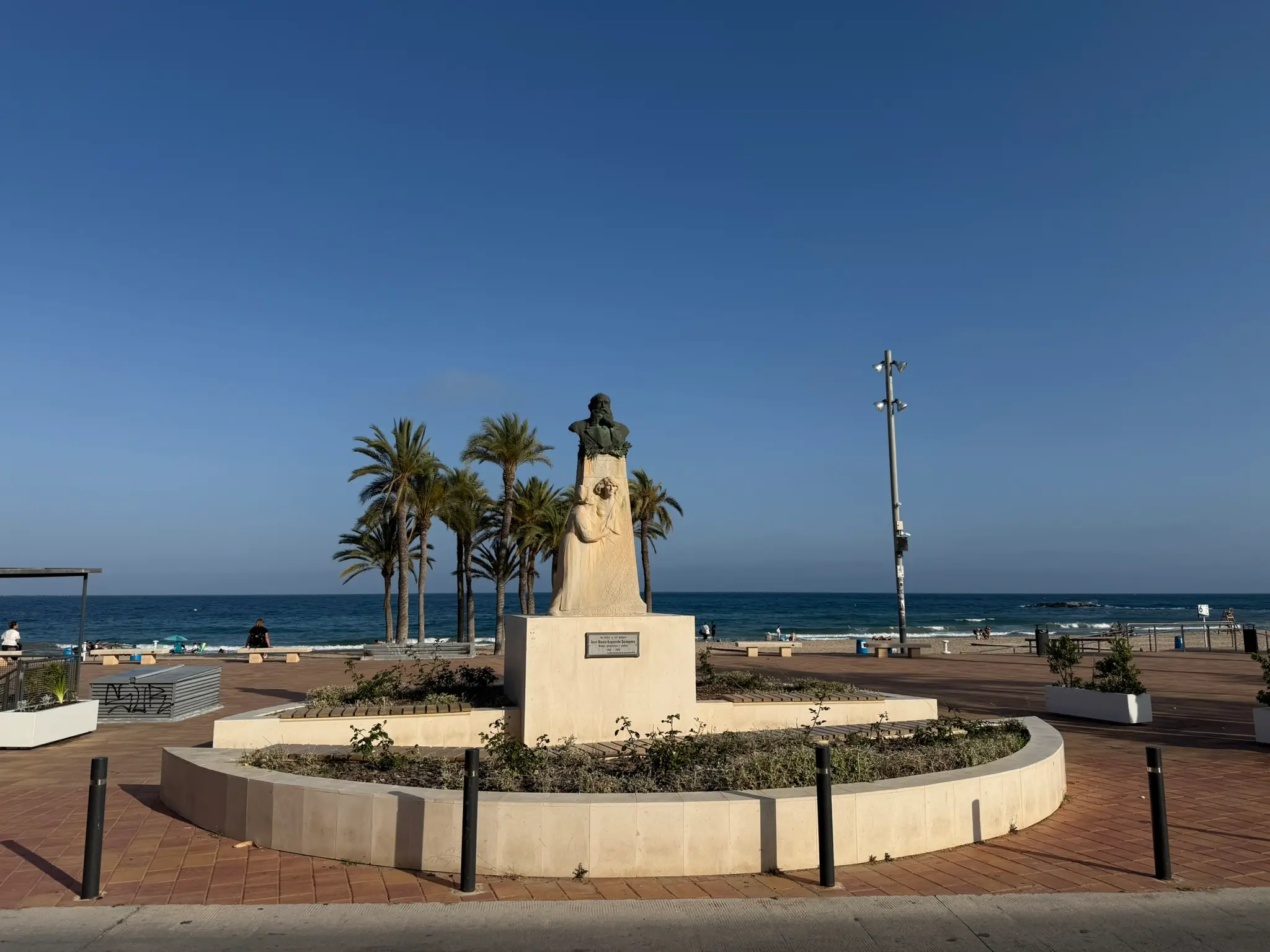The Jose Maria Esquerdo monument on the Villajoyosa beach promenade — stone bust in circular planter with palm trees and Mediterranean in background