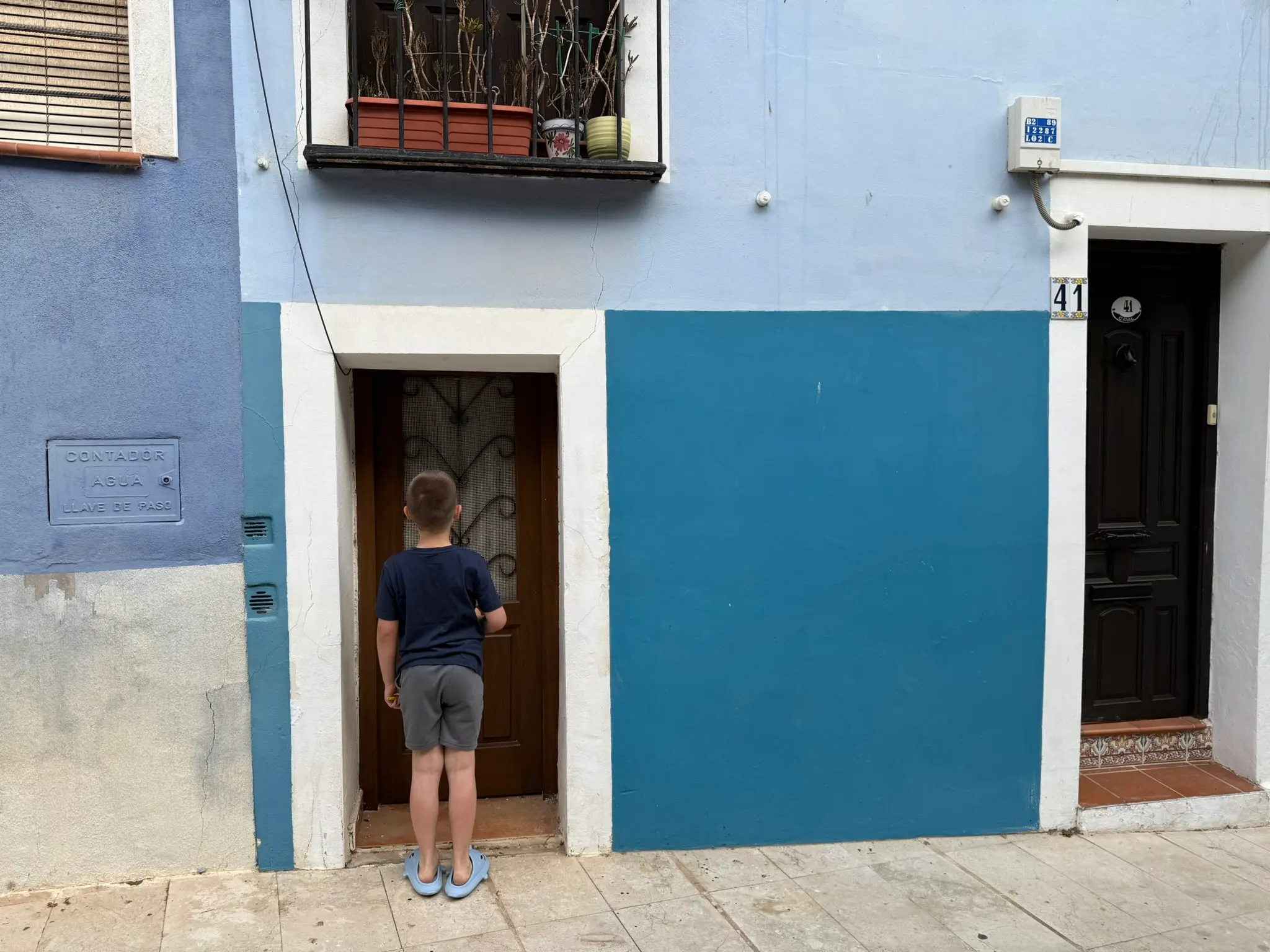 Traditional blue and light blue ground floor of house 41 in Villajoyosa — child in dark blue t-shirt facing wooden entrance door with ornamental ironwork