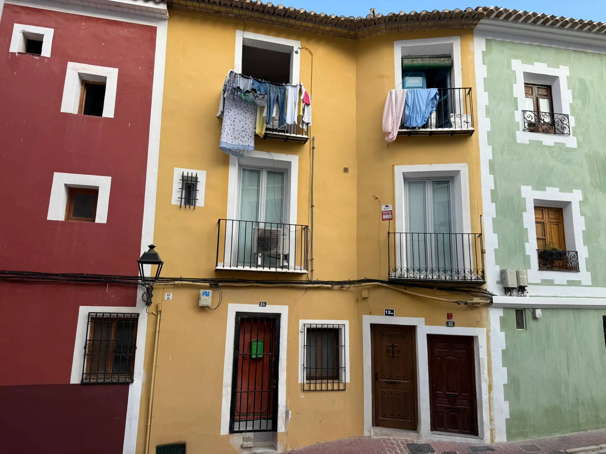 Brightly painted residential buildings in Villajoyosa — yellow corner house 12/14 with balconies and laundry drying, flanked by red and green facades