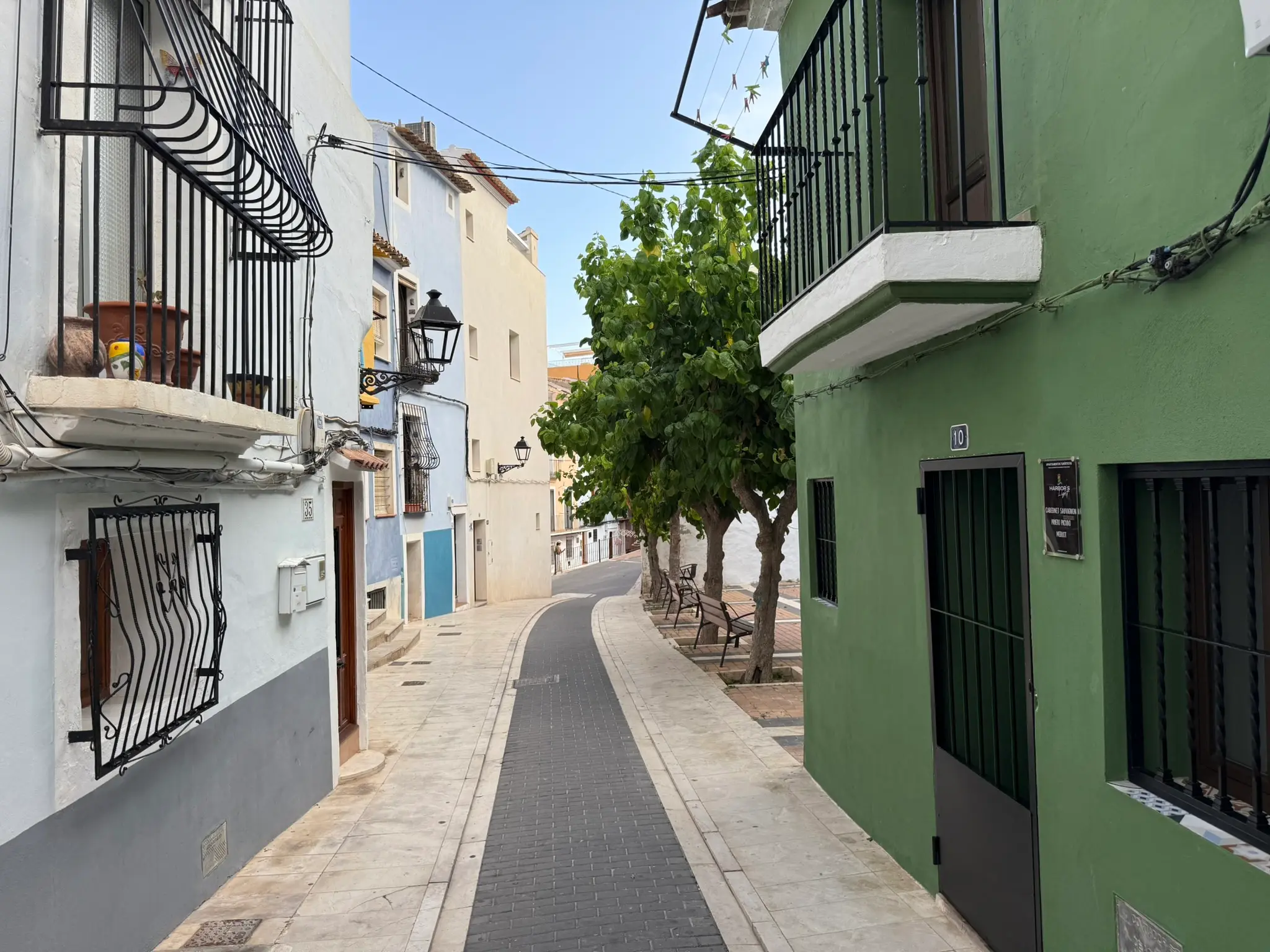 Scenic perspective of a pedestrian street in Villajoyosa — bright blue and orange buildings with wooden doors and balcony with potted plants, shady trees and benches