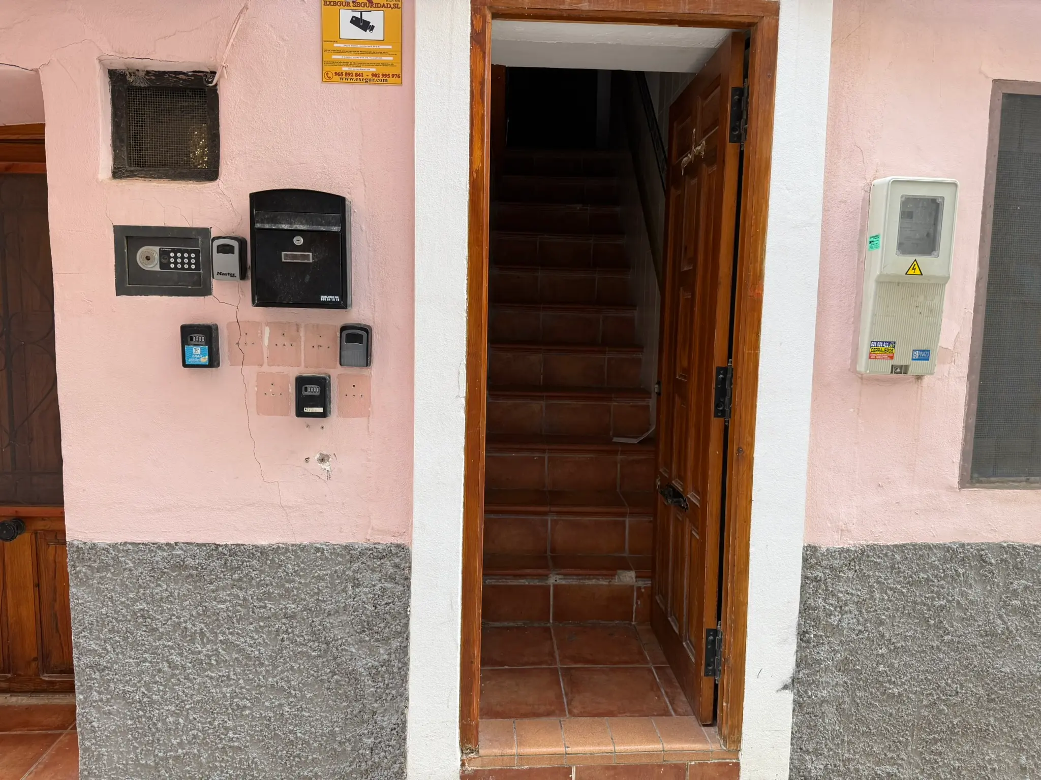 Open wooden entrance door to traditional holiday apartment in Villajoyosa — pink facade with key lockboxes, mailbox, electricity meter, and alarm sign