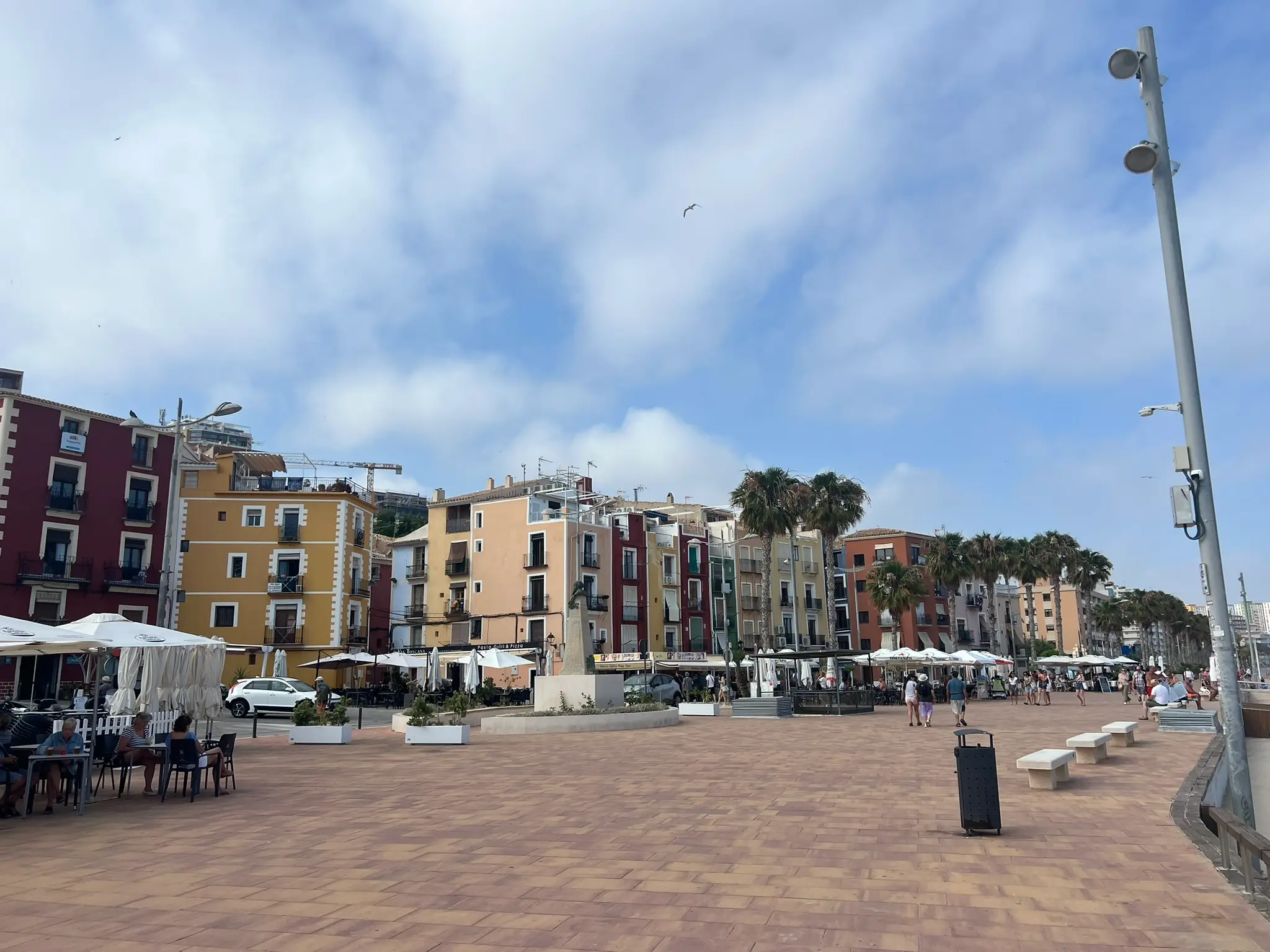 Wide view of bustling Paseo Marítimo beach promenade in Villajoyosa — outdoor restaurant terraces under white umbrellas, public benches, palm trees, colourful apartment buildings backdrop