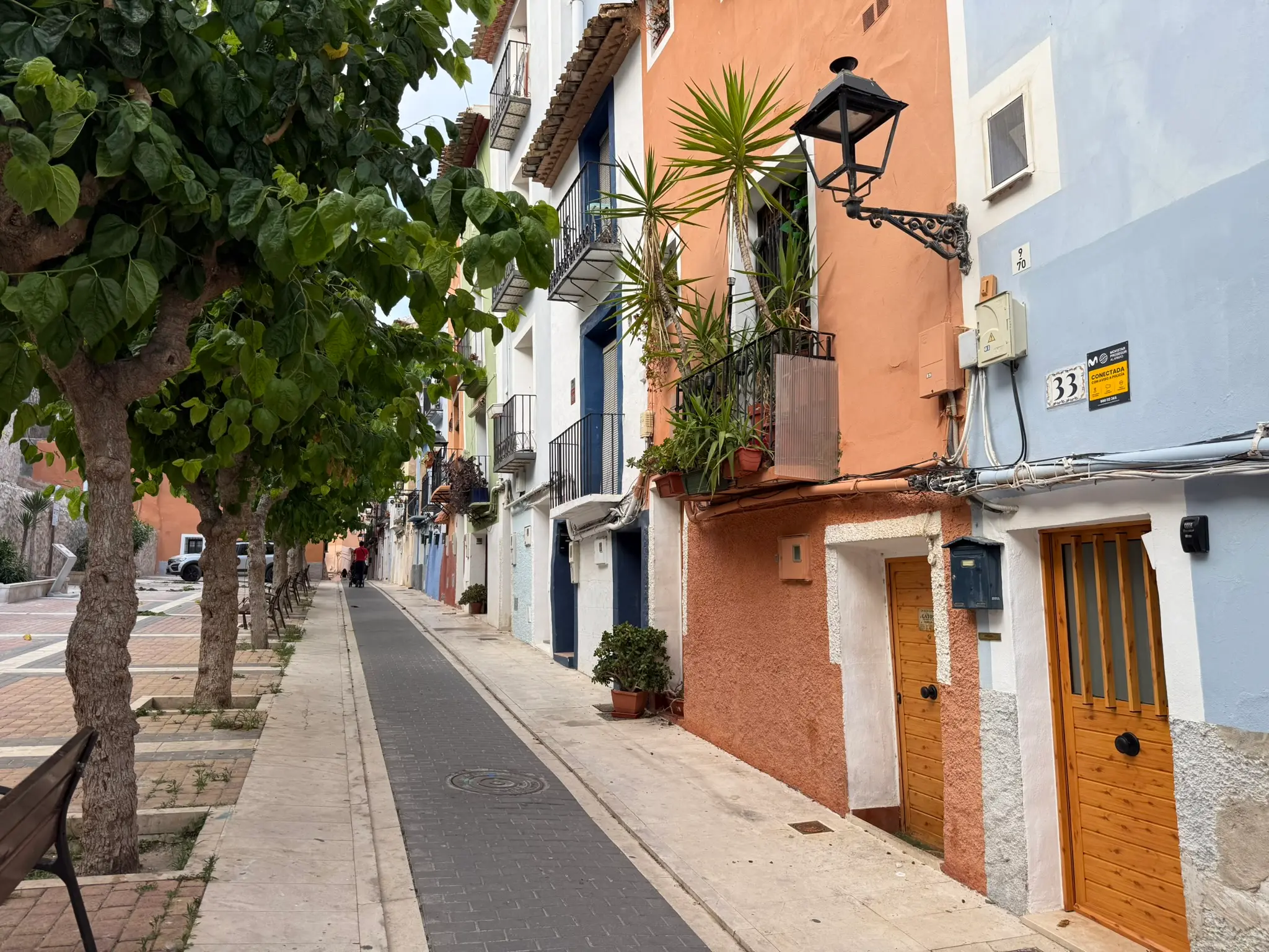 Paved pedestrian alley curving through Villajoyosa old town — dark green building with house number 10, light blue house with iron window grates, trees and benches